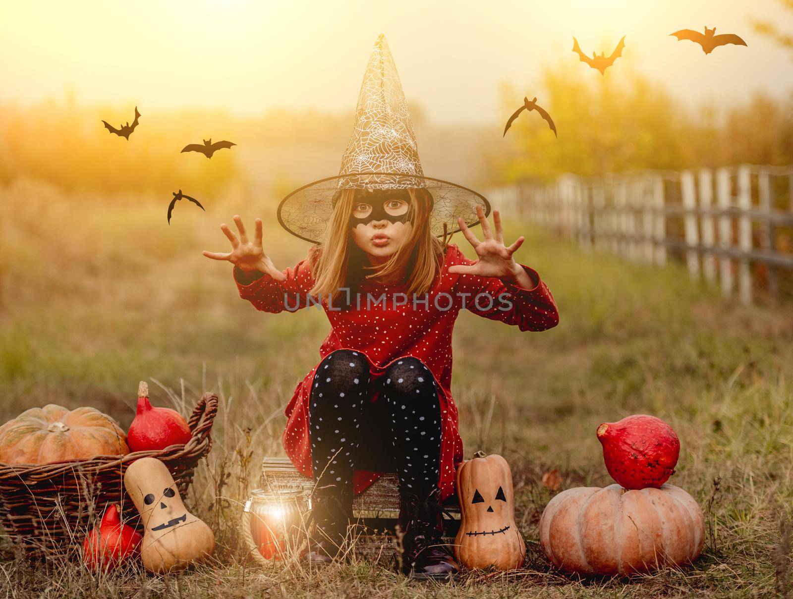 Little girl in halloween costume sitting next to pumpkins while frightening at camera on autumn nature