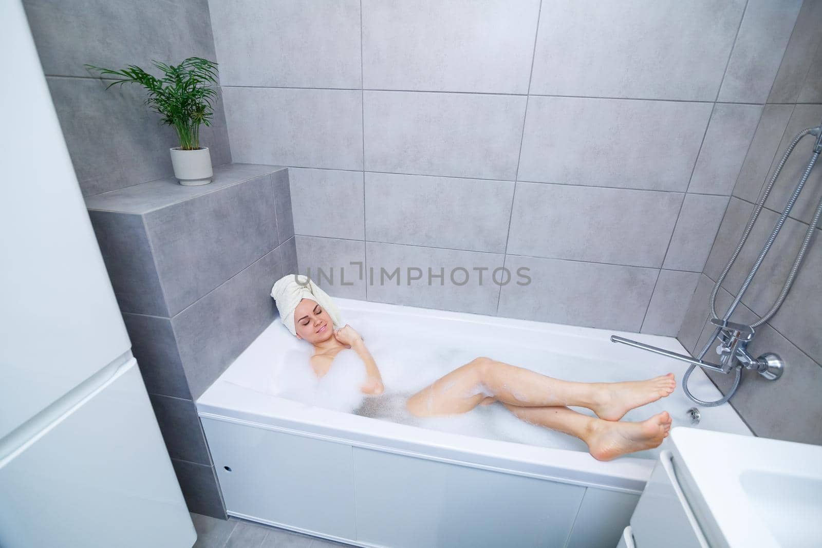 Girl woman with a white towel on her head lies in the bathroom in a white bath. In the hands of a lot of muddy soap. Full whirlpool foam. Relax after a hard day. Spa relaxing treatment