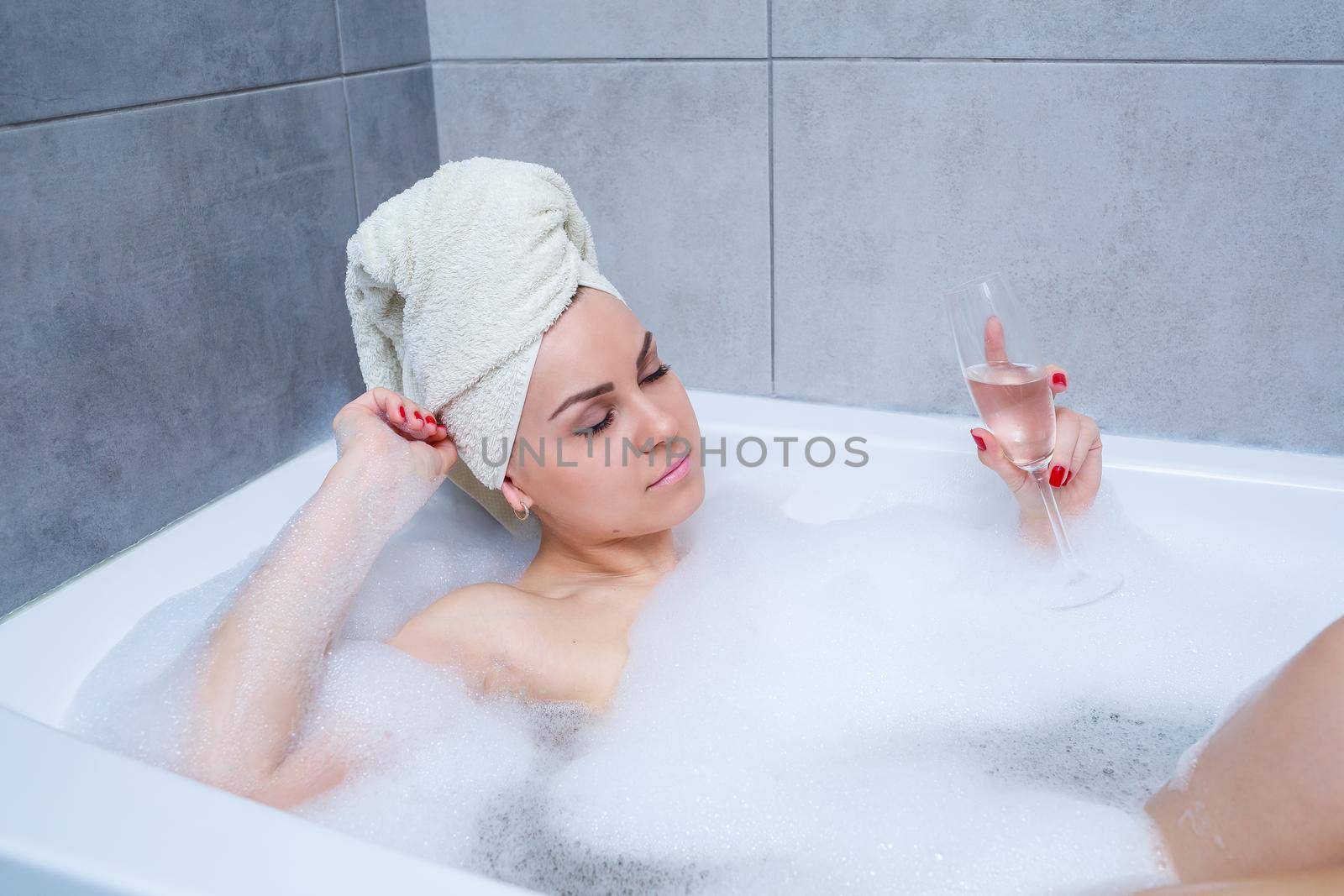Girl woman with a white towel on her head lies with a glass of champagne in a white bathtub. In her hands a lot of pee soap. Full whirlpool foam. Relax after a hard day. Spa relaxing procedure
