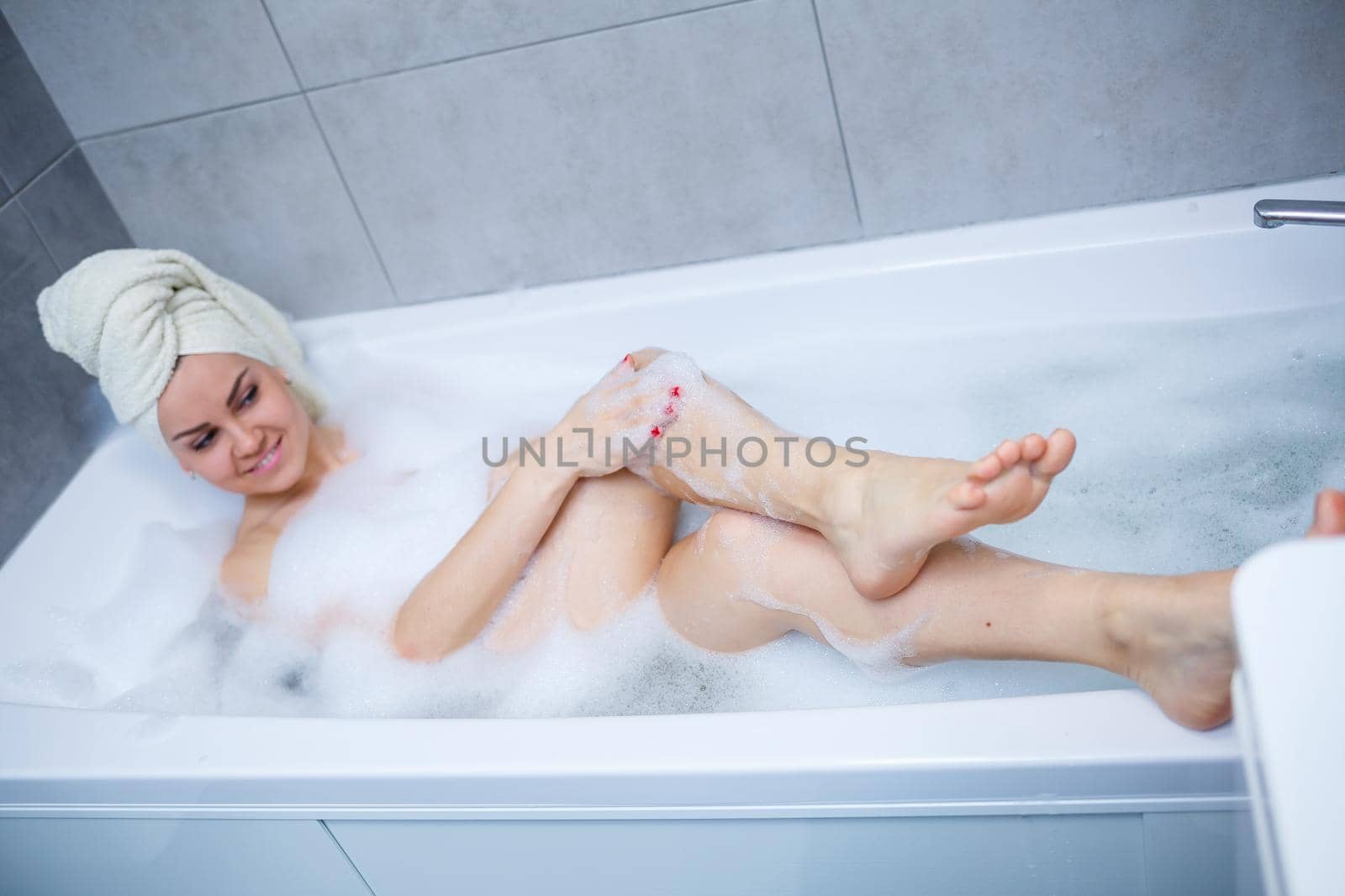 Girl woman with a white towel on her head lies in the bathroom in a white bath. In the hands of a lot of muddy soap. Full whirlpool foam. Relax after a hard day. Spa relaxing treatment