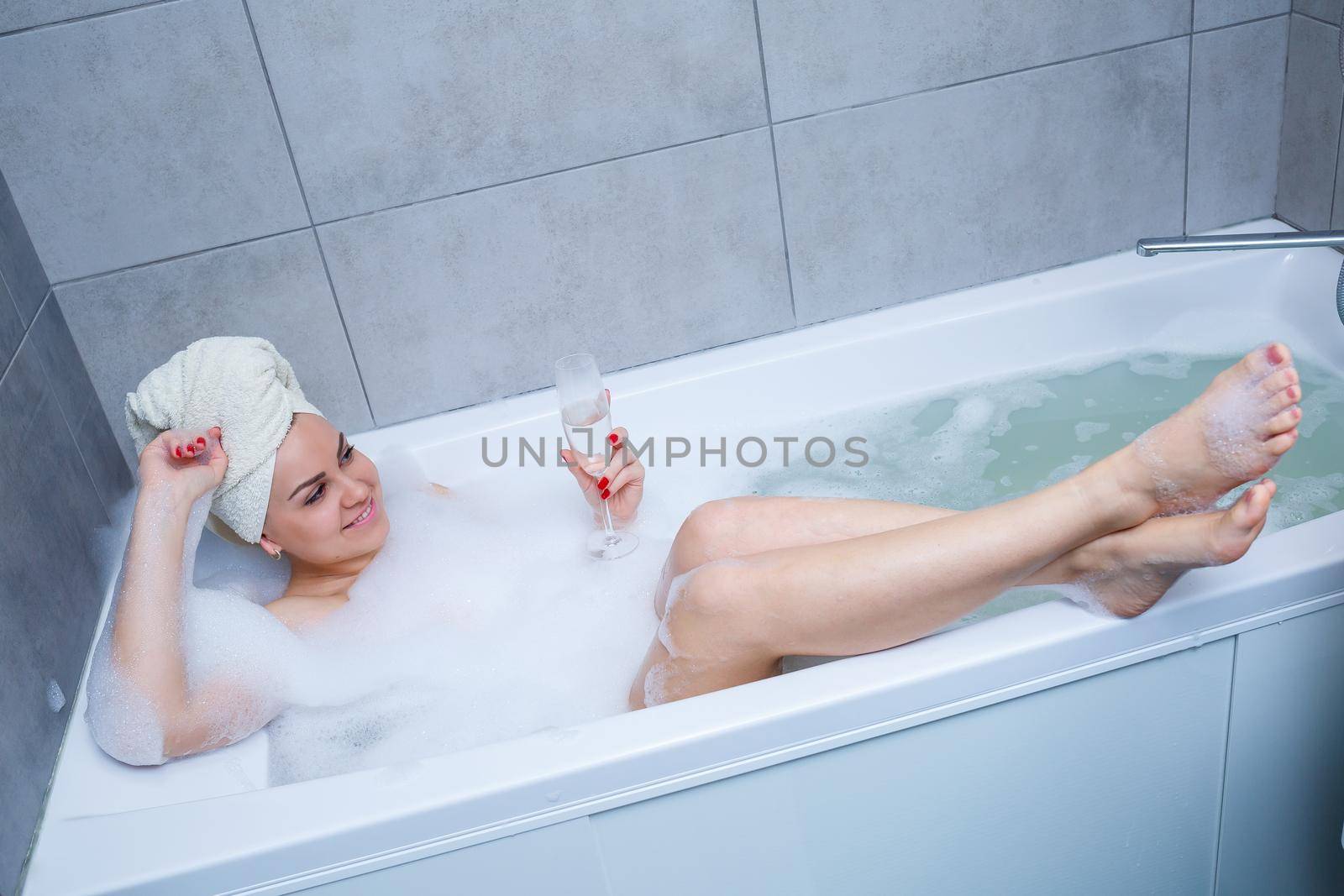 Girl woman with a white towel on her head lies with a glass of champagne in a white bathtub. In her hands a lot of pee soap. Full whirlpool foam. Relax after a hard day. Spa relaxing procedure