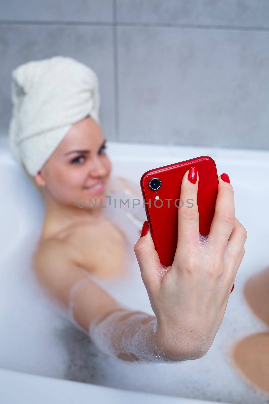 Girl woman with a towel on her head lies with a glass of champagne in a white bathtub and takes a selfie on the phone. In her hands a lot of drunk soap. Relax after a hard day. Spa relaxing procedure