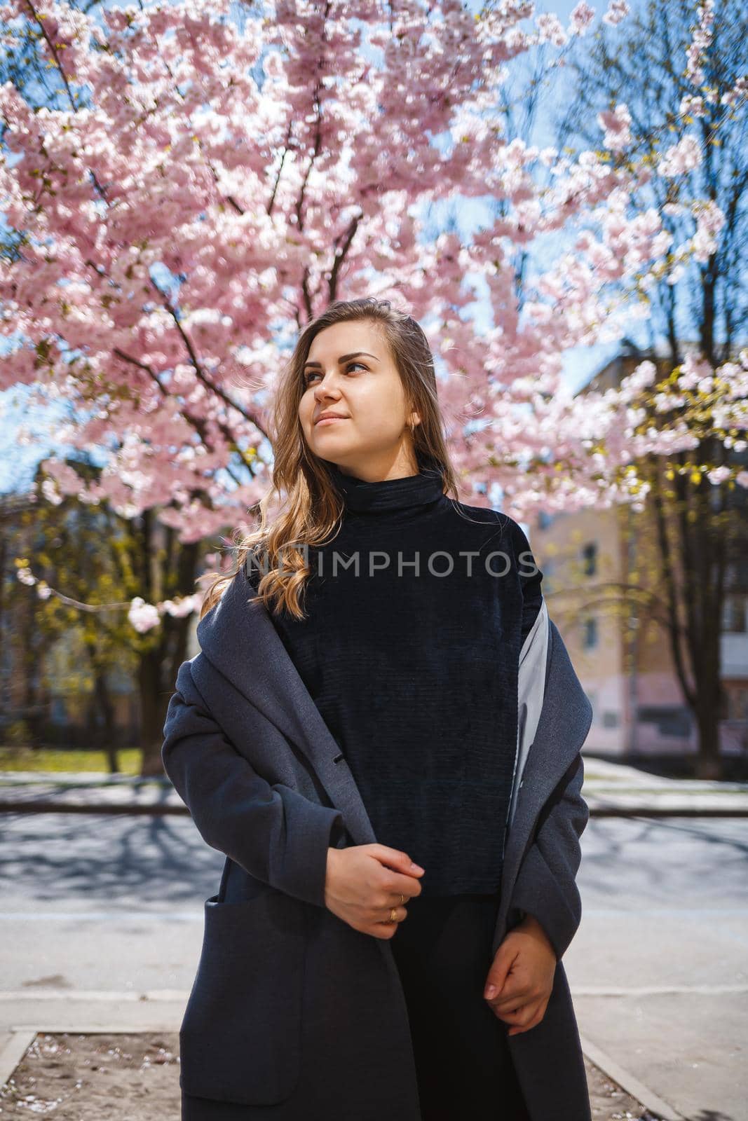A young slender female model with long wavy hair and, dressed in a gray coat, sneakers, stands on the street near a flowering shrub with beautiful pink flowers in the background and poses.