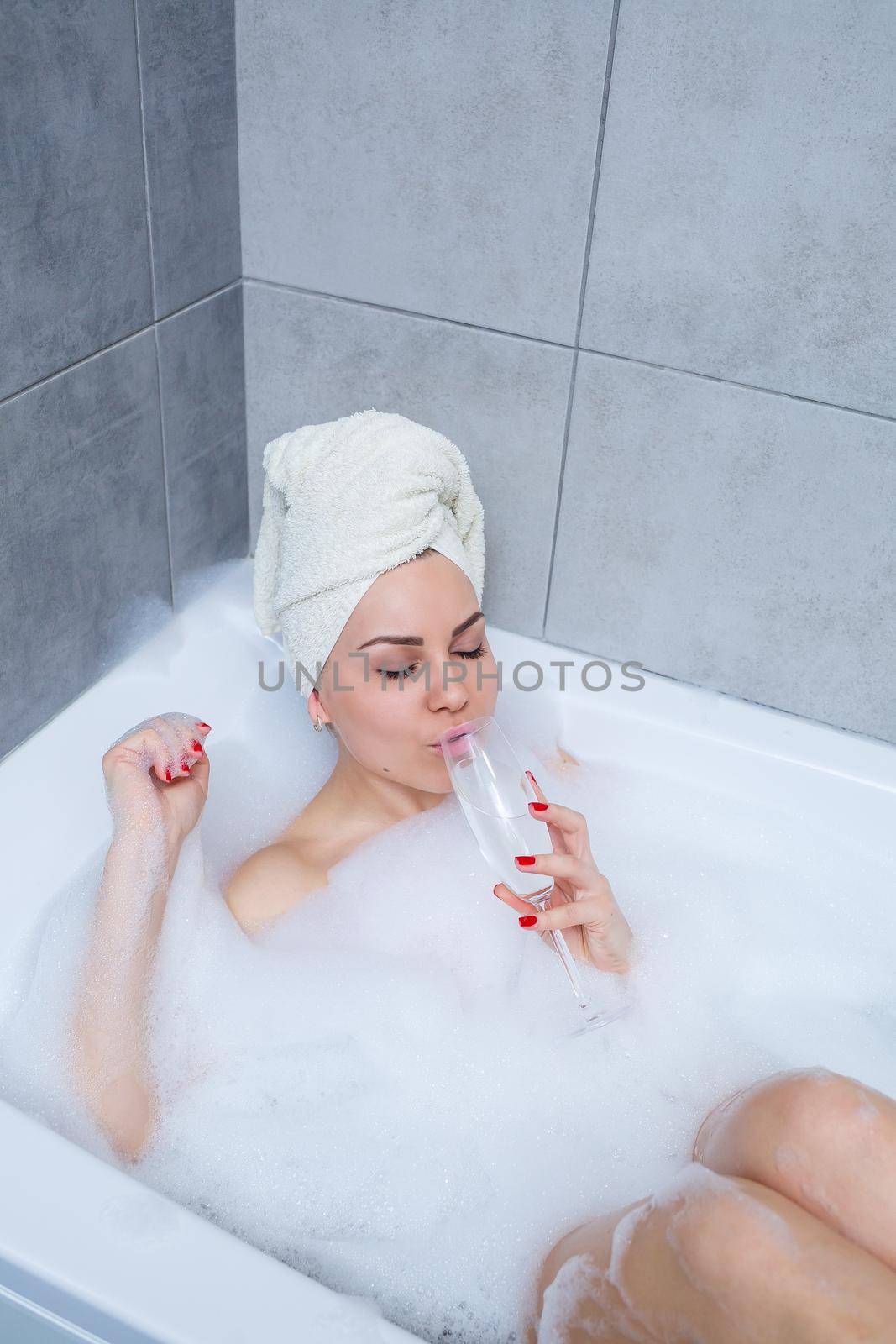 Girl woman with a white towel on her head lies with a glass of champagne in a white bathtub. In her hands a lot of pee soap. Full whirlpool foam. Relax after a hard day. Spa relaxing procedure