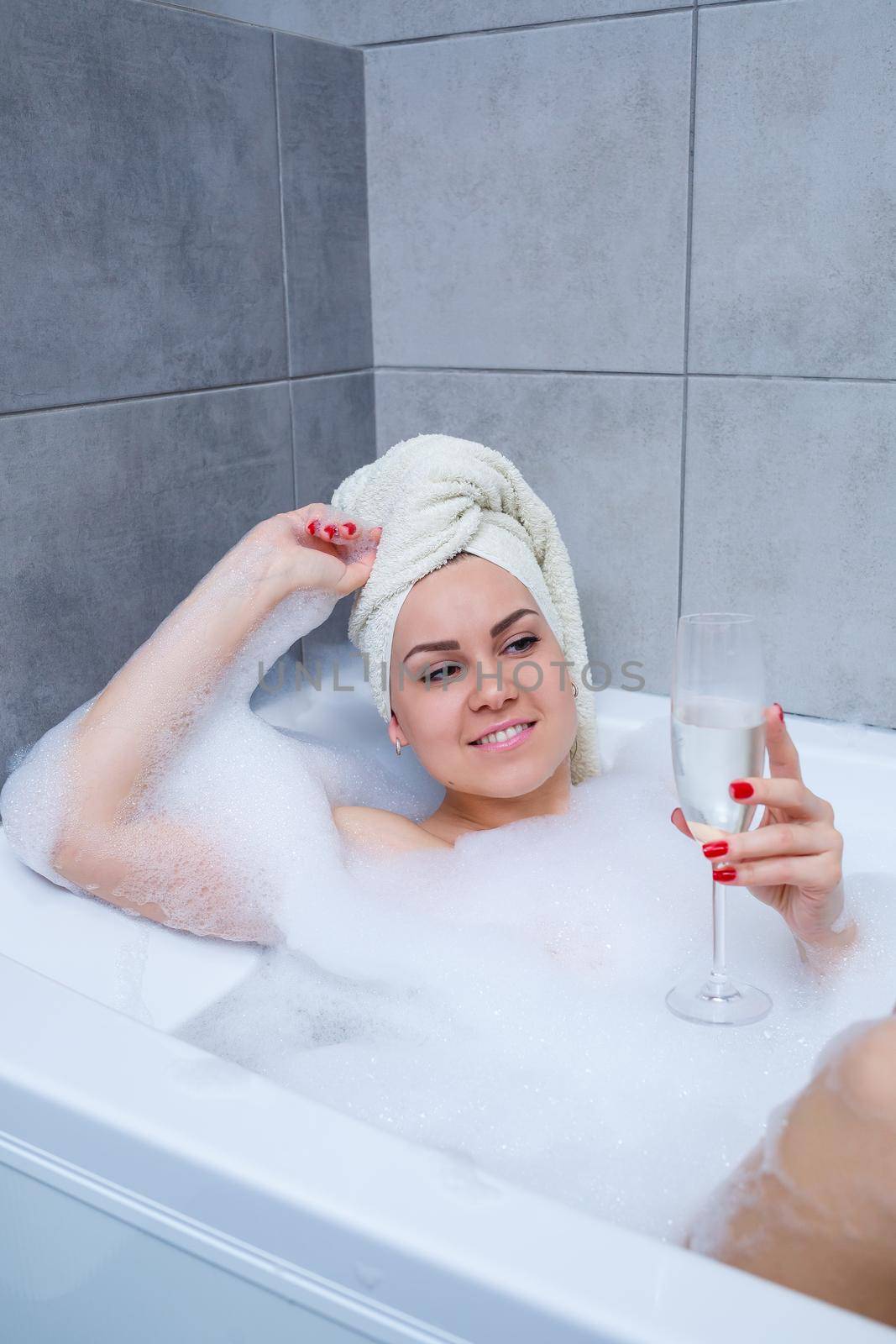 Girl woman with a white towel on her head lies with a glass of champagne in a white bathtub. In her hands a lot of pee soap. Full whirlpool foam. Relax after a hard day. Spa relaxing procedure