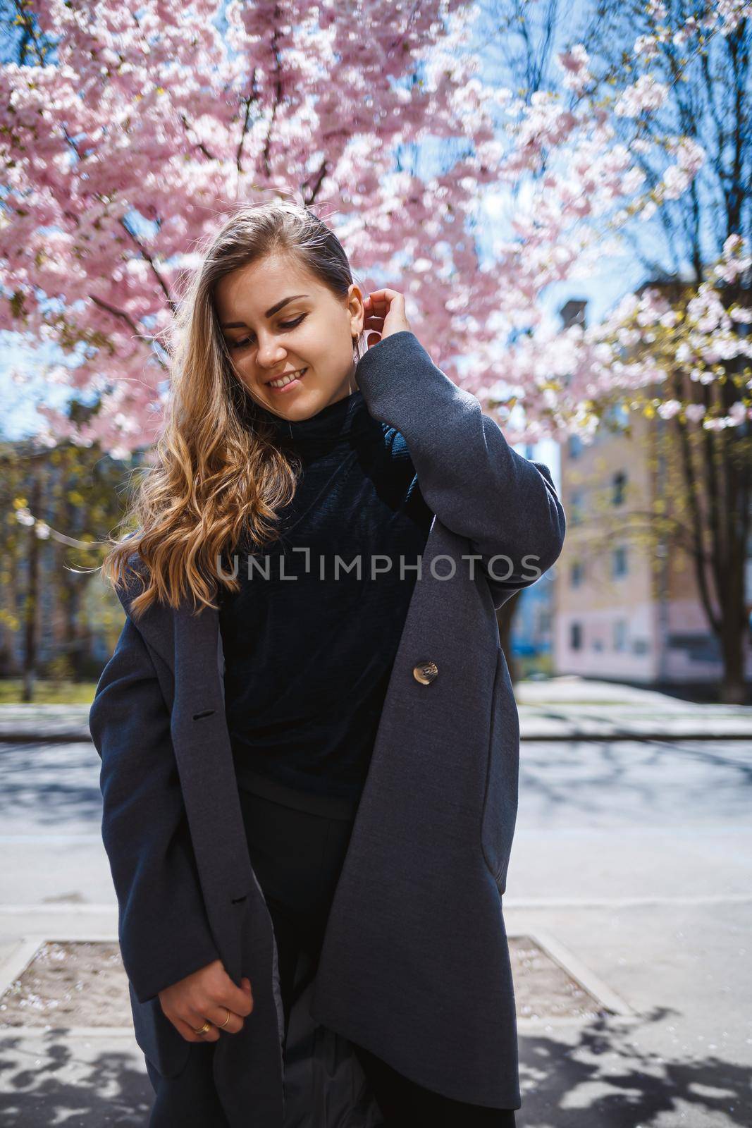 A young slender female model with long wavy hair and, dressed in a gray coat, sneakers, stands on the street near a flowering shrub with beautiful pink flowers in the background and poses.