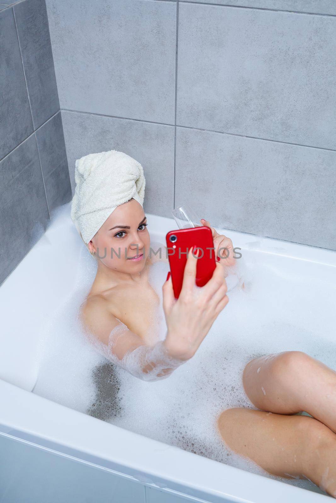 Girl woman with a towel on her head lies with a glass of champagne in a white bathtub and takes a selfie on the phone. In her hands a lot of drunk soap. Relax after a hard day. Spa relaxing procedure