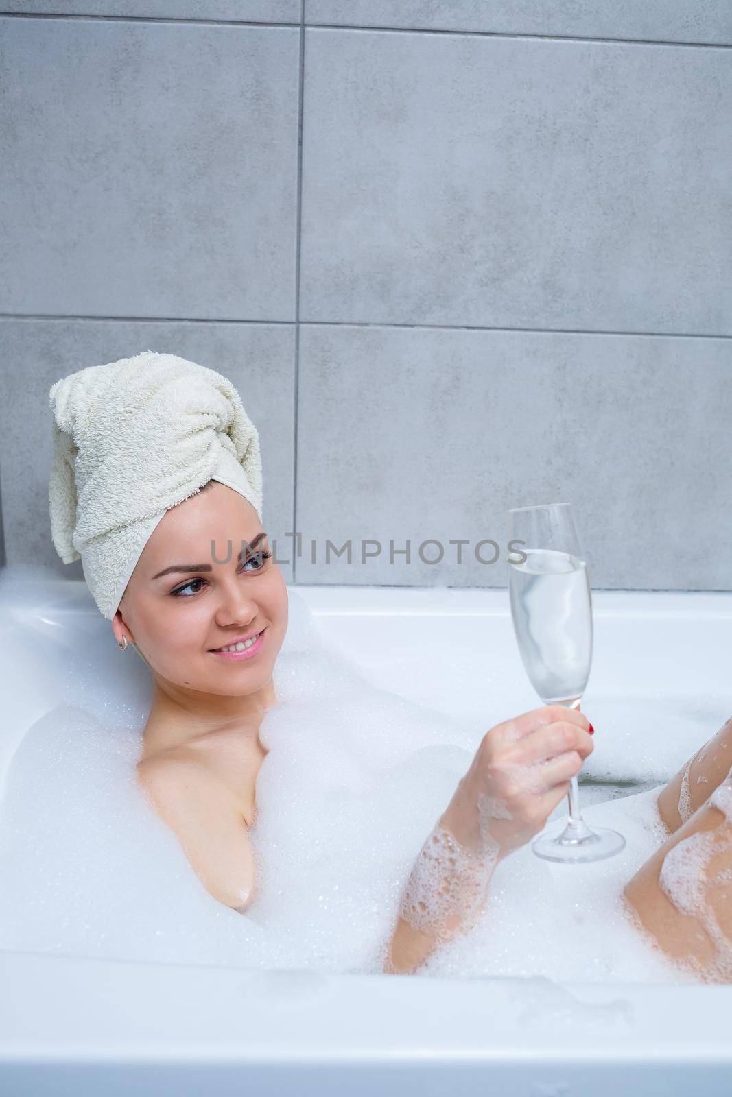 Girl woman with a white towel on her head lies with a glass of champagne in a white bathtub. In her hands a lot of pee soap. Full whirlpool foam. Relax after a hard day. Spa relaxing procedure