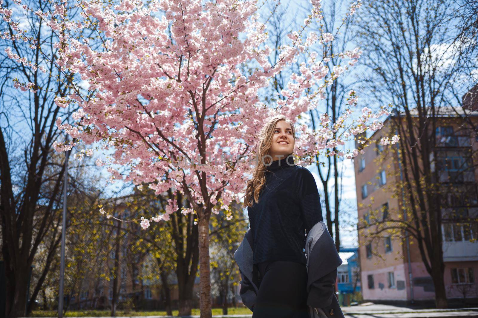 A young slender female model with long wavy hair and, dressed in a gray coat, sneakers, stands on the street near a flowering shrub with beautiful pink flowers in the background and poses.