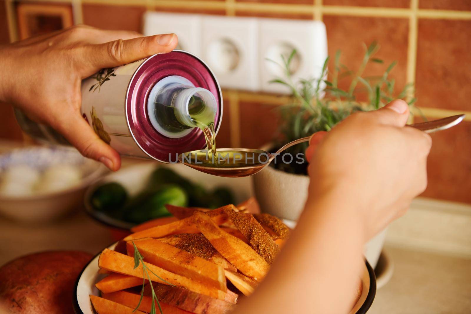 Selective focus. Close-up of chef's hands pouring organic olive oil on a spoon, and sprinkling it over organic sweet potato slices while preparing lunch in the home kitchen