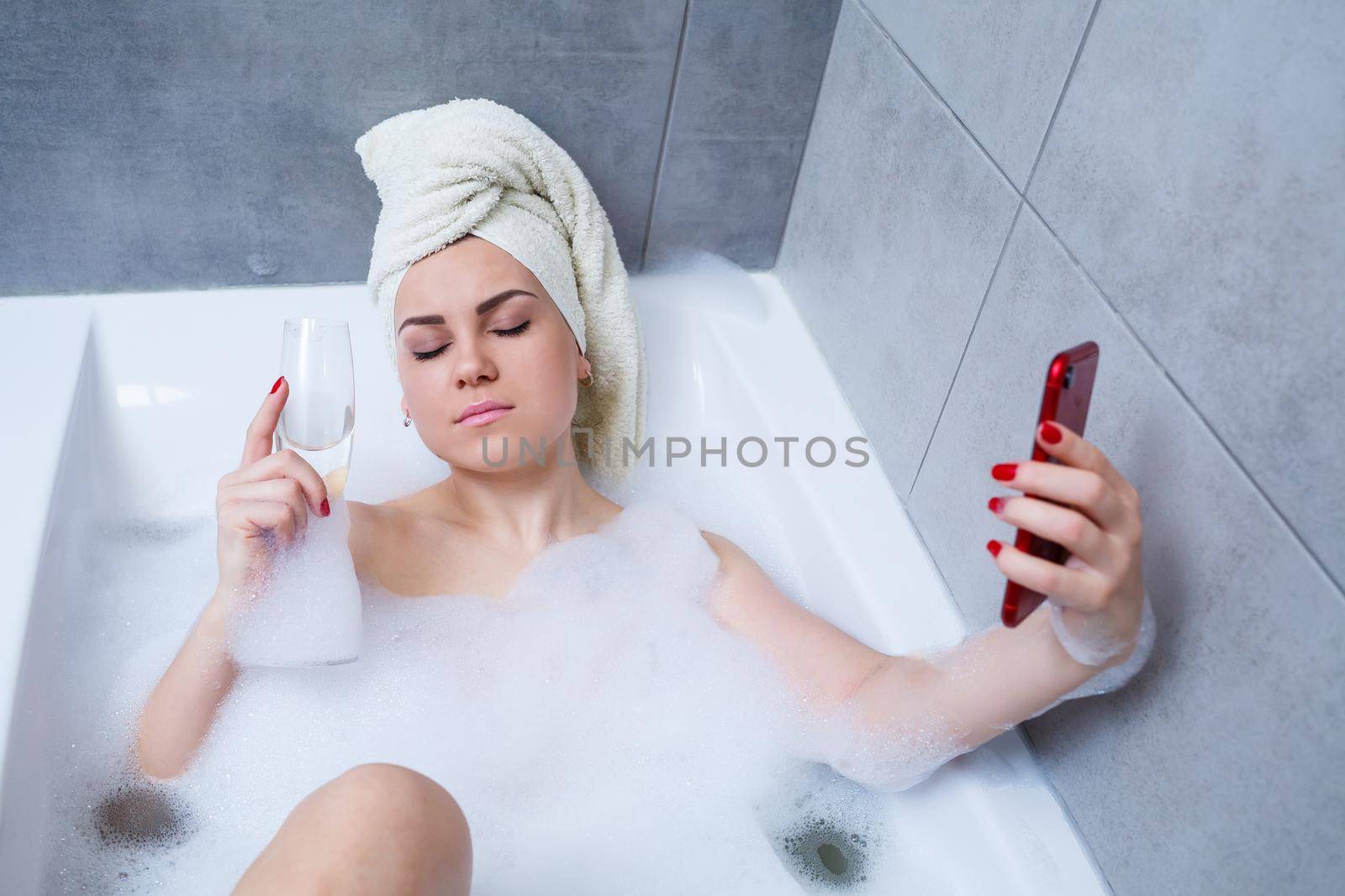 Girl woman with a towel on her head lies with a glass of champagne in a white bathtub and takes a selfie on the phone. In her hands a lot of drunk soap. Relax after a hard day. Spa relaxing procedure
