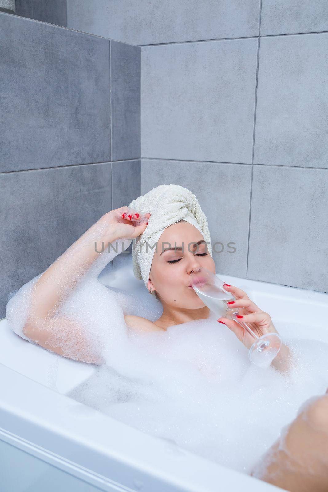 Girl woman with a white towel on her head lies with a glass of champagne in a white bathtub. In her hands a lot of pee soap. Full whirlpool foam. Relax after a hard day. Spa relaxing procedure