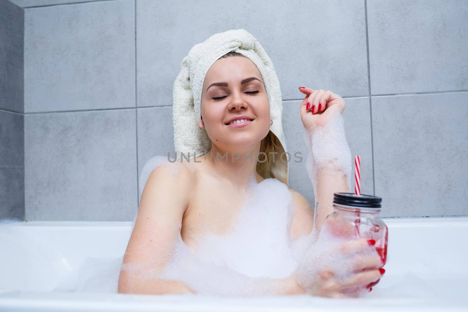Beautiful young woman with a towel on her head drinking a cocktail takes a bath at home. Relax after a hard day. The spa is a relaxing procedure.