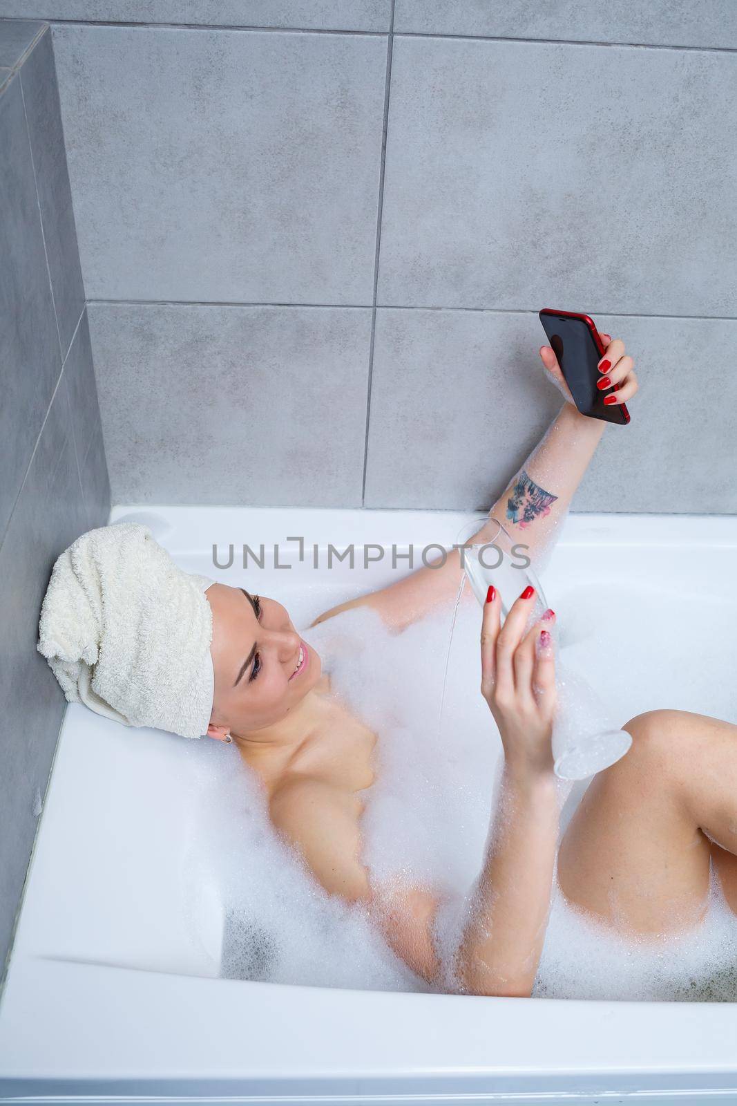 Girl woman with a towel on her head lies with a glass of champagne in a white bathtub and takes a selfie on the phone. In her hands a lot of drunk soap. Relax after a hard day. Spa relaxing procedure