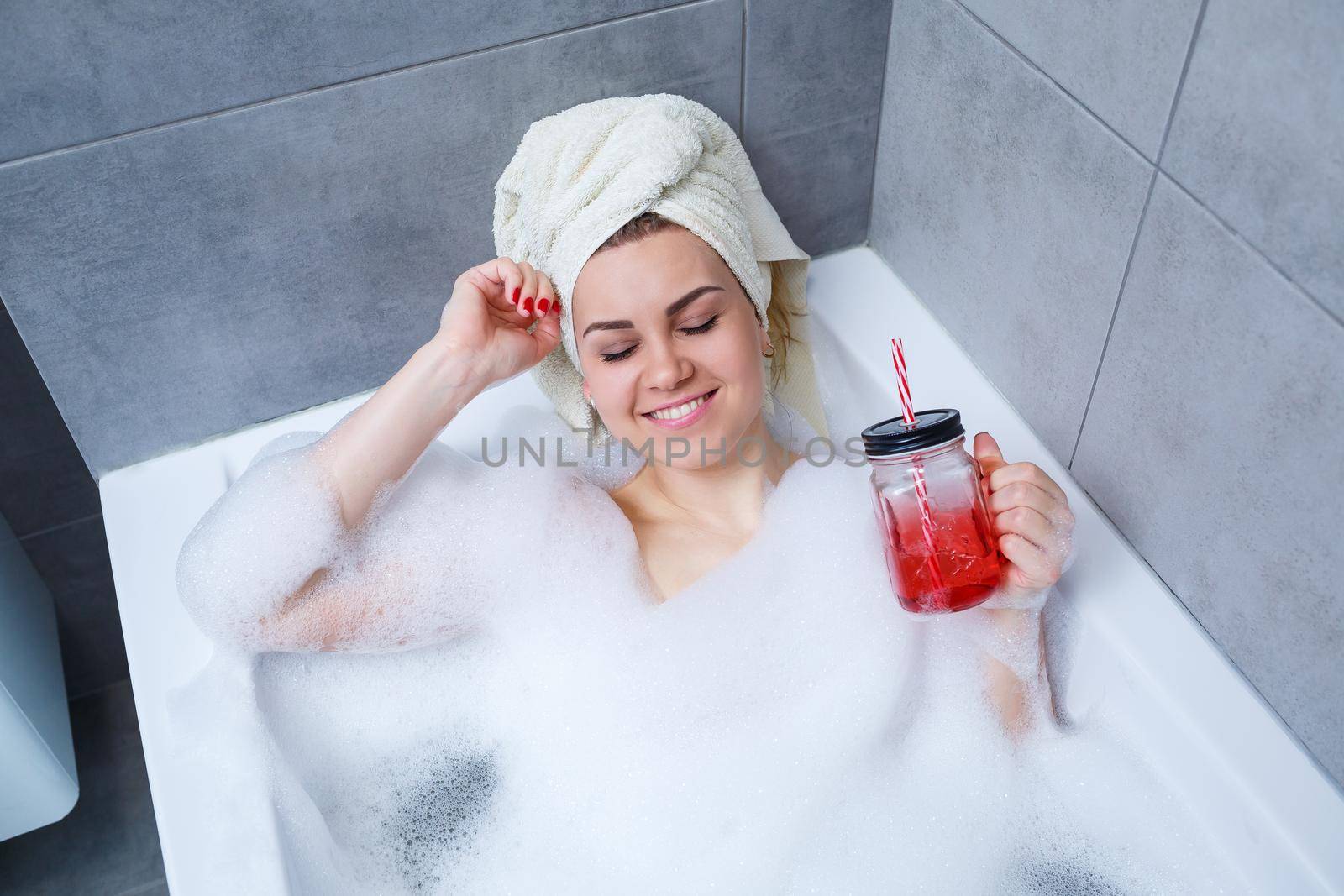 Beautiful young woman with a towel on her head drinking a cocktail takes a bath at home. Relax after a hard day. The spa is a relaxing procedure.
