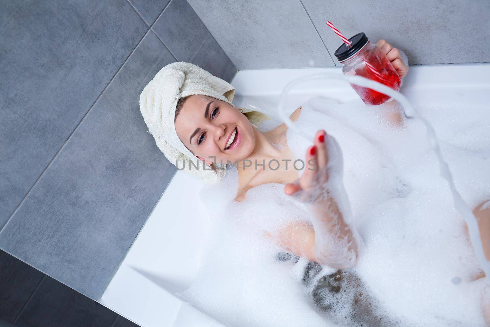 Beautiful young woman with a towel on her head drinking a cocktail takes a bath at home. Relax after a hard day. The spa is a relaxing procedure.