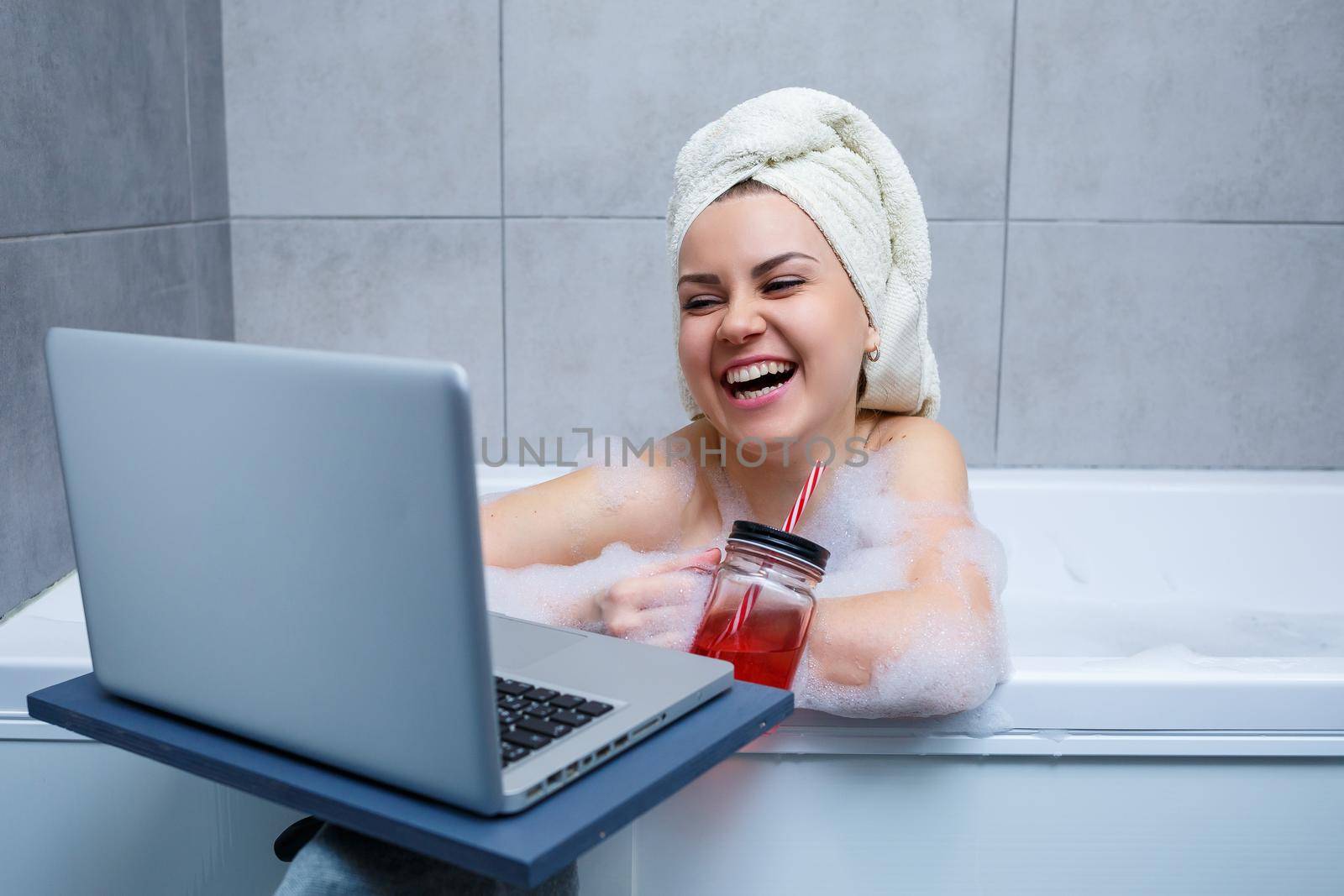 A young woman with a towel on her head drinks juice watching a movie on a laptop while sitting in a bath in a beauty salon. Relax in the bathroom without linen. Body care and relaxation concept.
