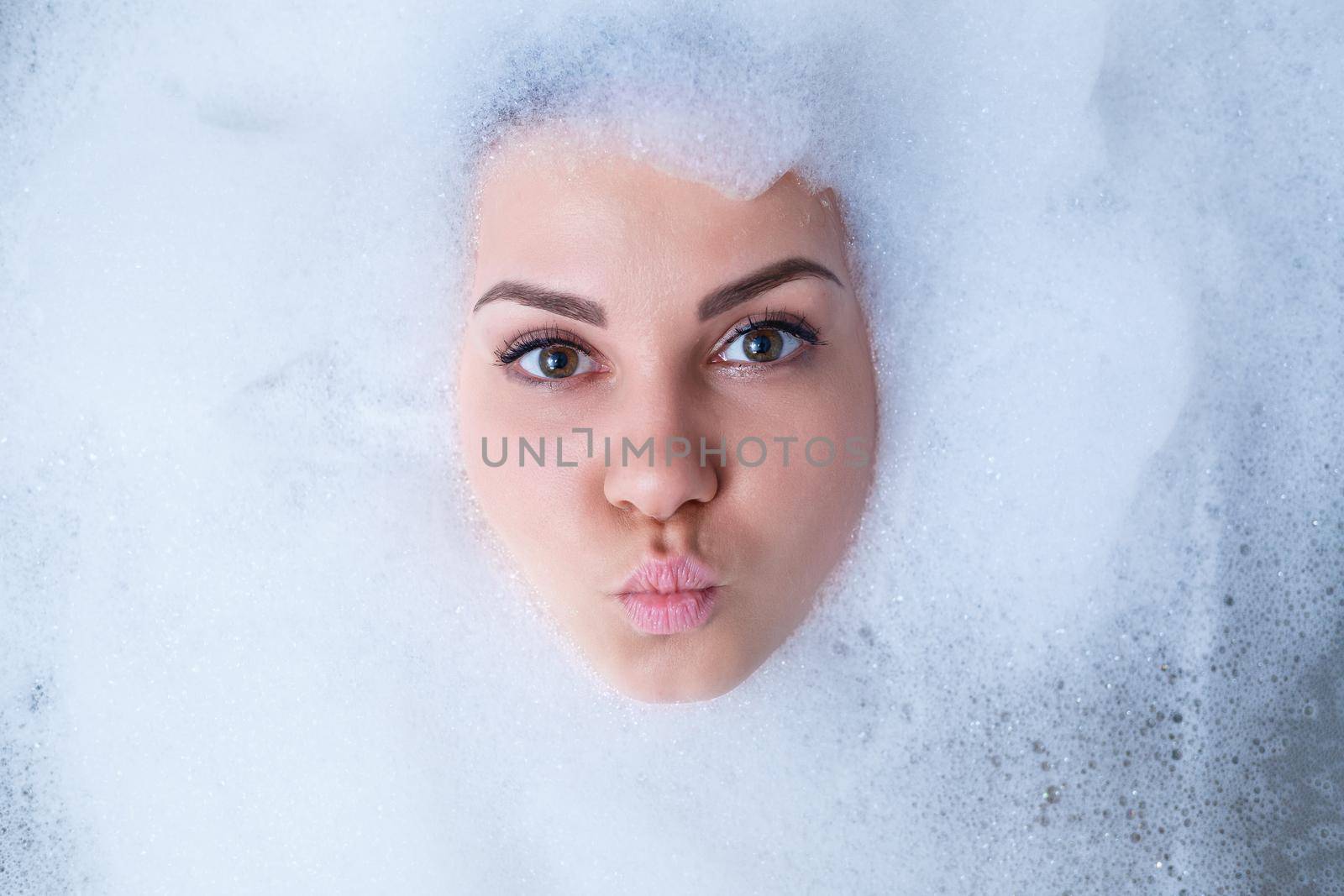 Closeup portrait of a girl in a bathtub and white foam around her face. Different emotions, grimaces, woman smile