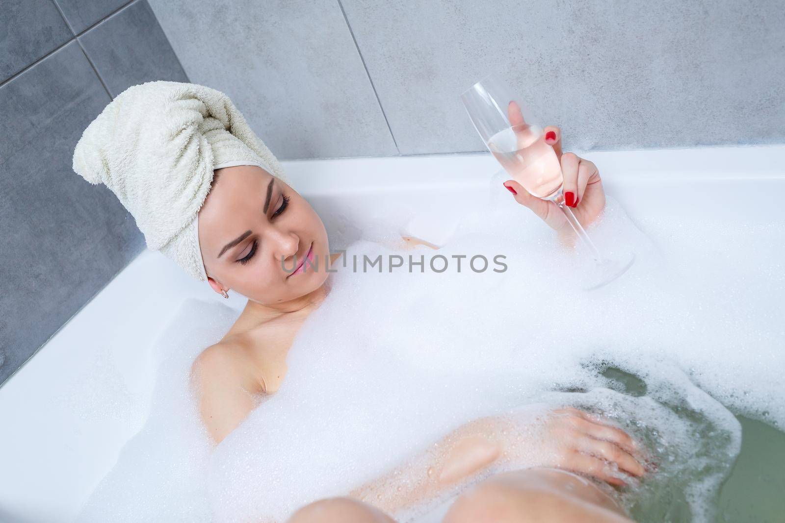 Girl woman with a white towel on her head lies with a glass of champagne in a white bathtub. In her hands a lot of pee soap. Full whirlpool foam. Relax after a hard day. Spa relaxing procedure
