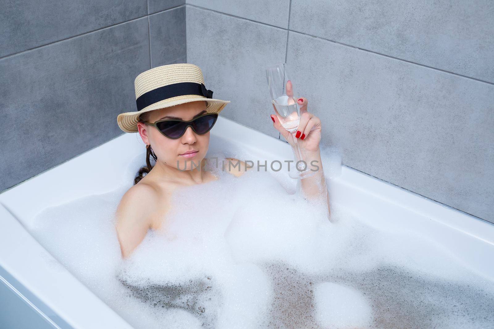 Beautiful young woman in a hat on her head and sunglasses drinking a cocktail takes a bath at home. Relax after a hard day. The spa is a relaxing procedure.