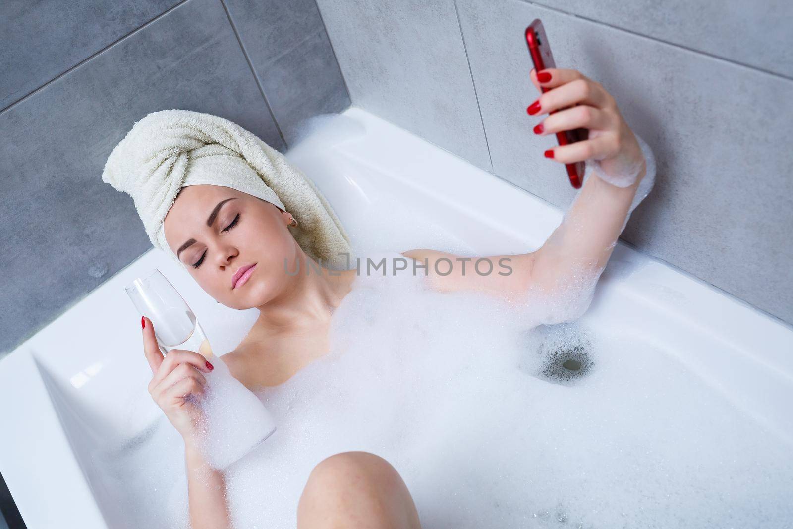 A beautiful woman with a towel on her head lies with a glass of champagne in a white bath and takes a selfie on the phone. Relax after a hard day. Relaxing spa treatment