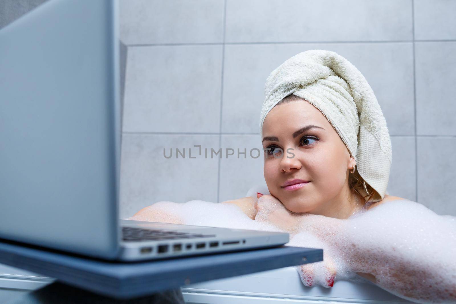 A young woman with a towel on her head is watching a movie on a laptop while sitting in a bath in a beauty salon. Relax in the bathroom without linen. Body care and relaxation concept.