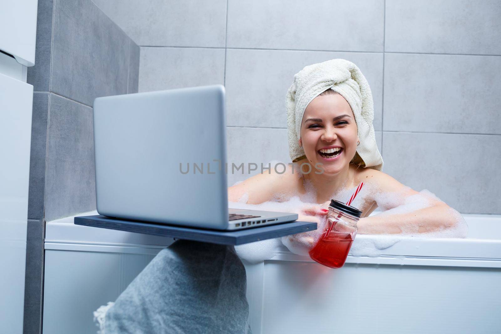 A young woman with a towel on her head drinks juice watching a movie on a laptop while sitting in a bath in a beauty salon. Relax in the bathroom without linen. Body care and relaxation concept.