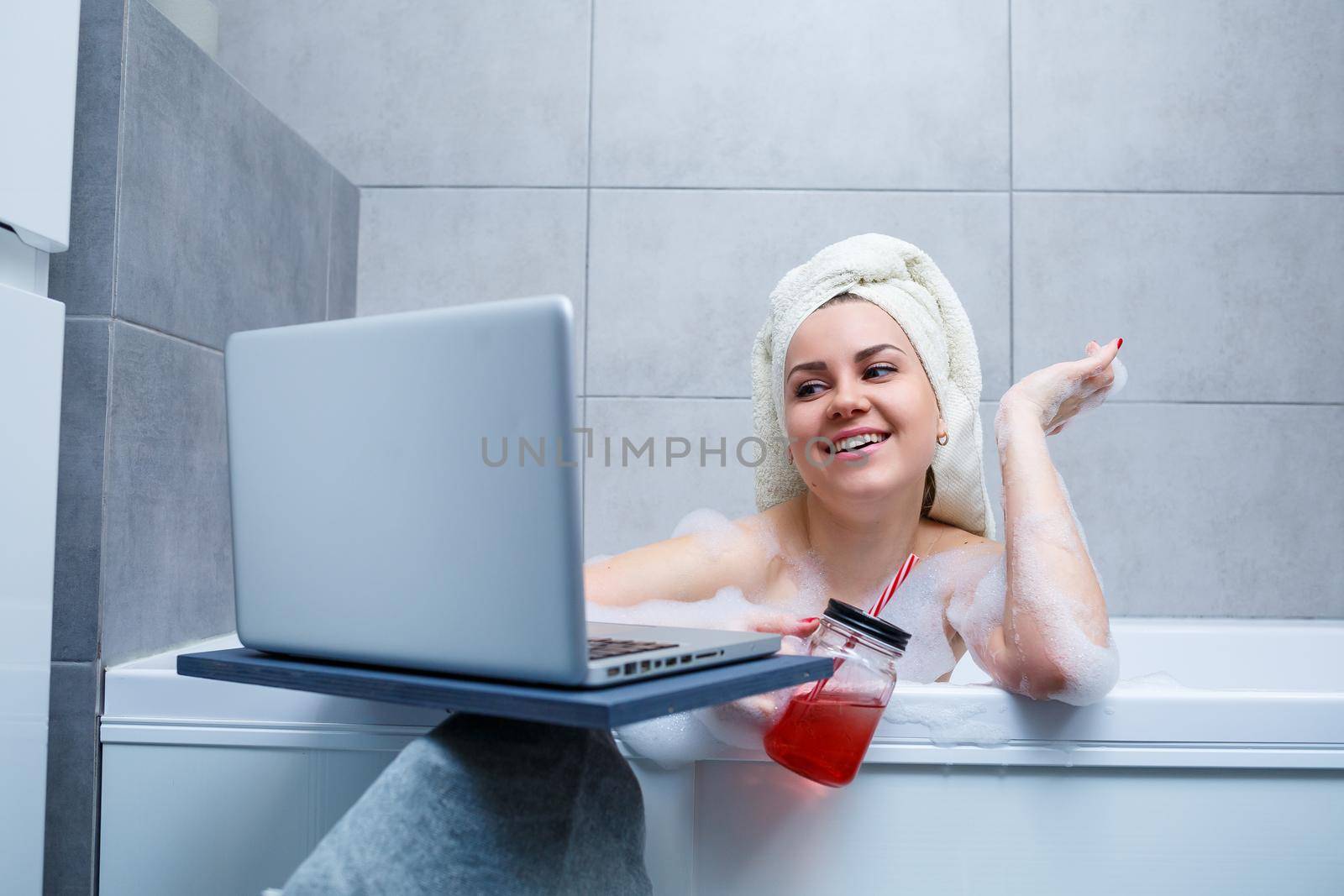 A young woman with a towel on her head drinks juice watching a movie on a laptop while sitting in a bath in a beauty salon. Relax in the bathroom without linen. Body care and relaxation concept.