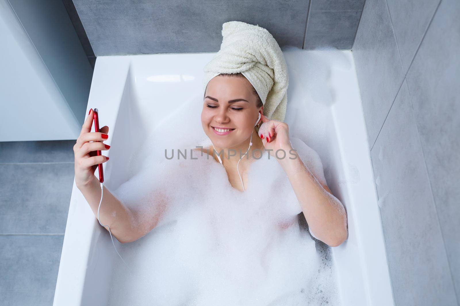 Beautiful young woman listening to music and singing while taking a bath at home. Relax after a hard day. Spa relaxing procedure.