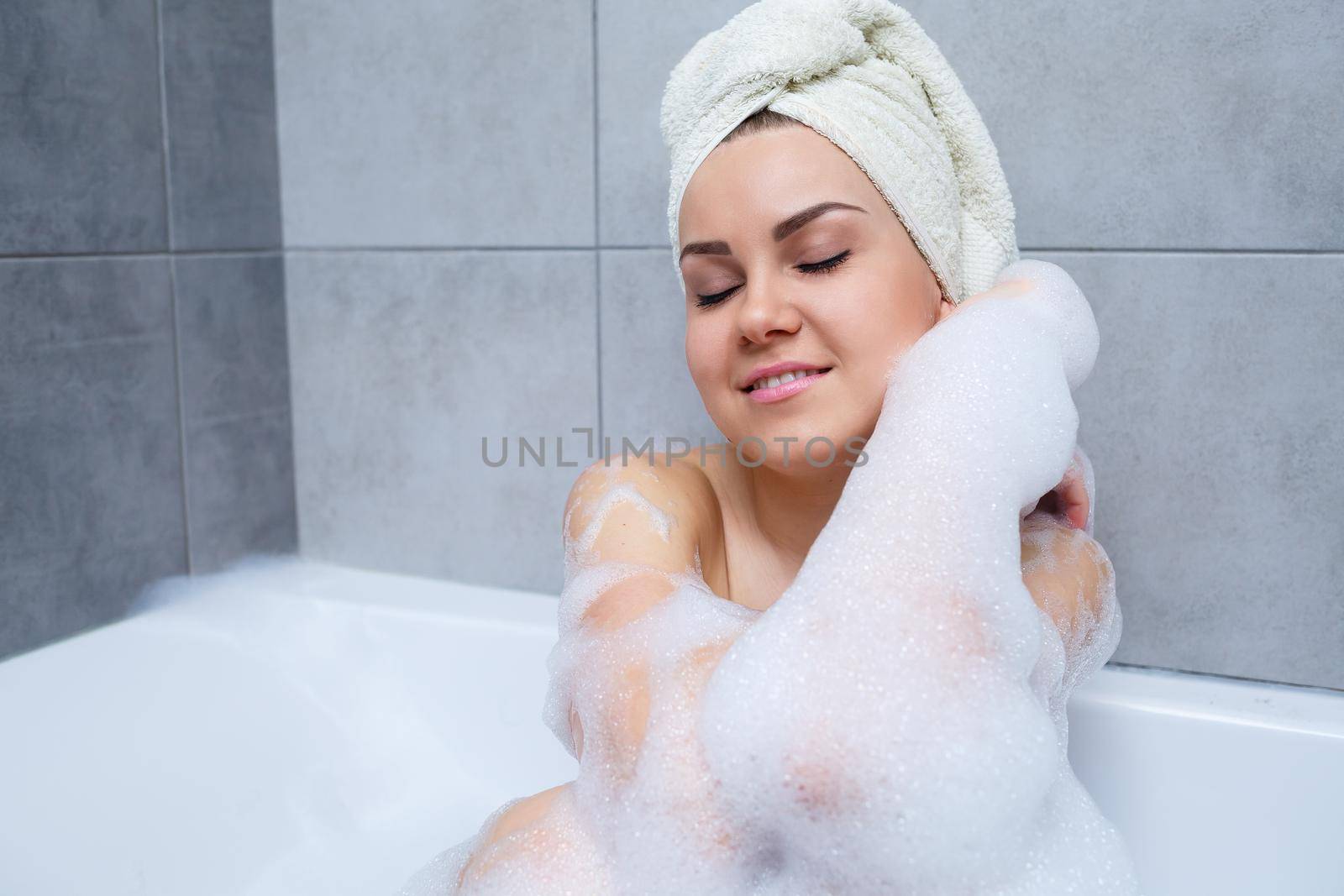 Girl woman with a white towel on her head lies in the bathroom in a white bath. Foam for around. Relax after a hard day. Spa relaxing treatment