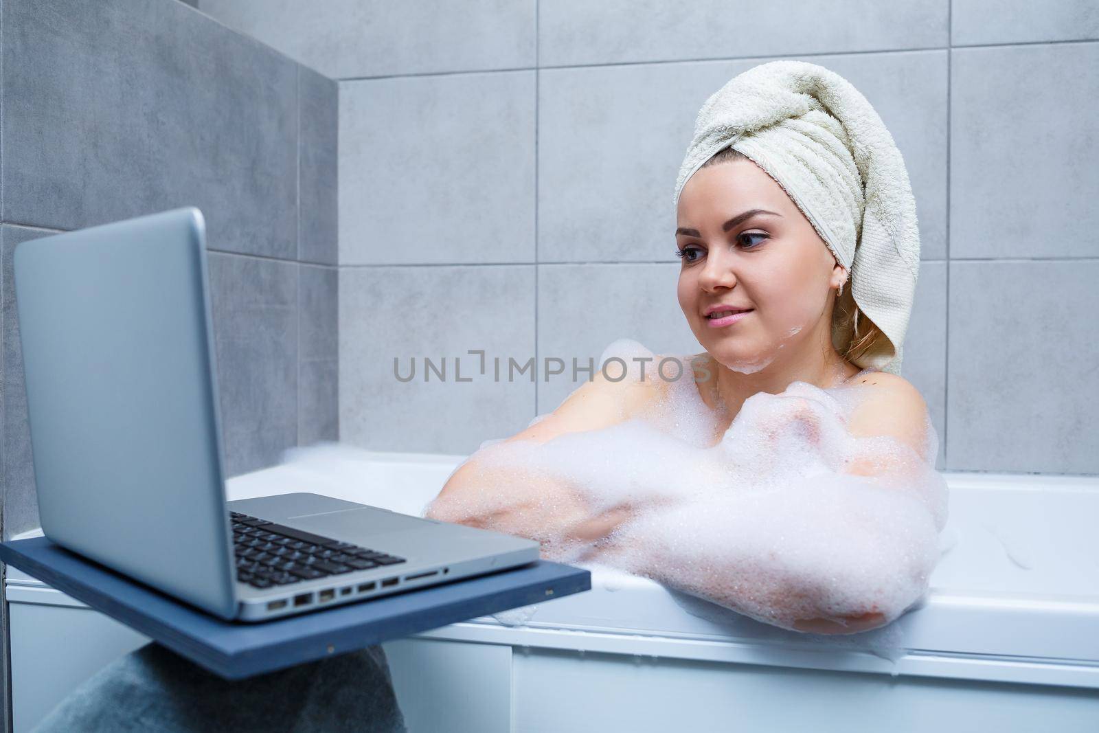 A young woman with a towel on her head is watching a movie on a laptop while sitting in a bath in a beauty salon. Relax in the bathroom without linen. Body care and relaxation concept.