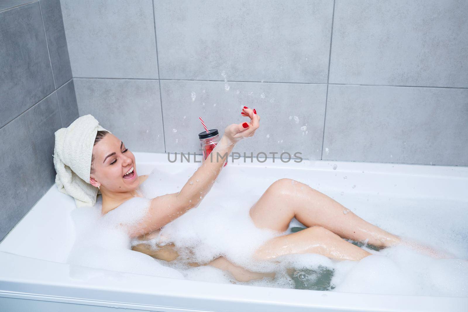 Beautiful young woman with a towel on her head, drinking a cocktail, takes a bath at home. Relax after a hard day.