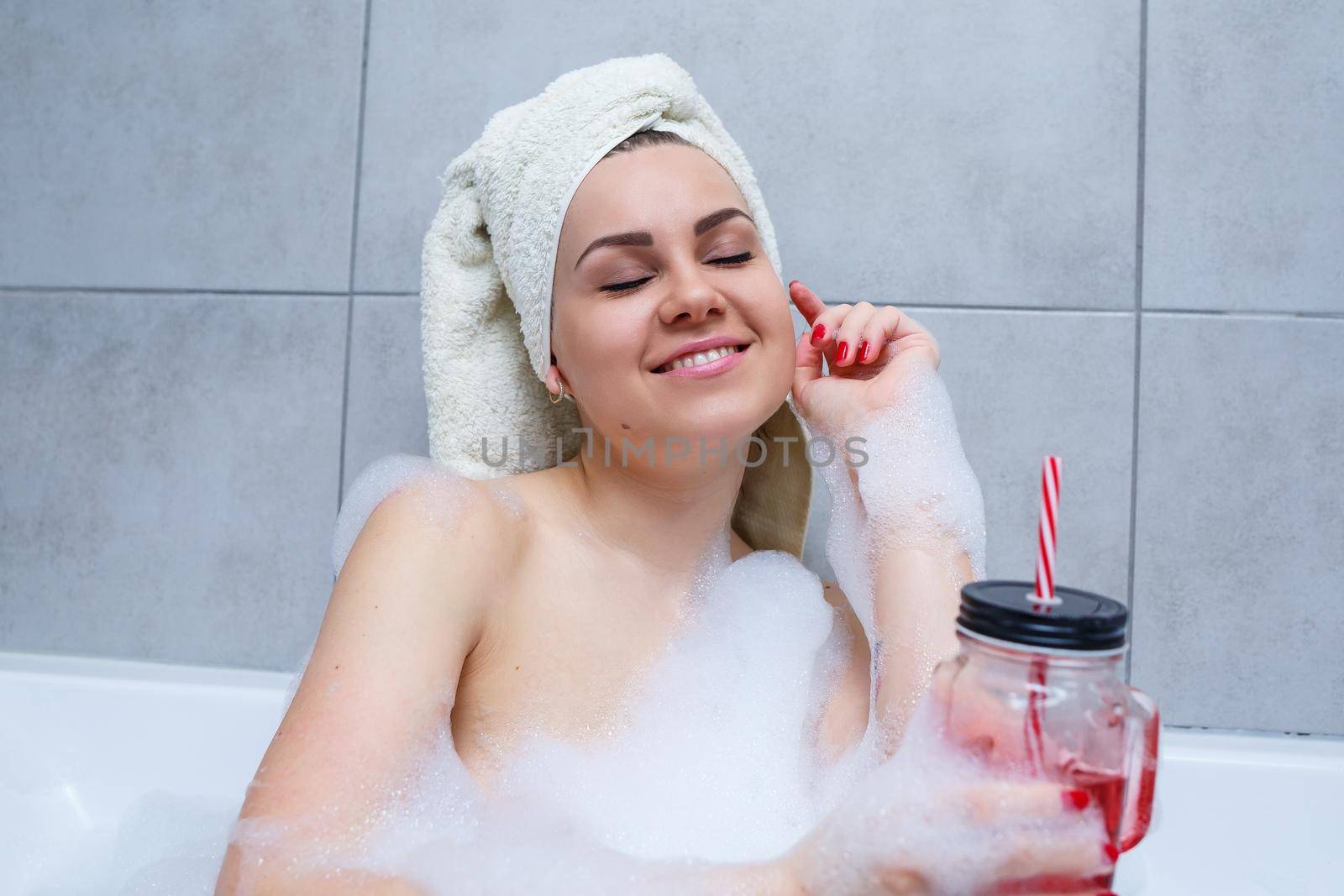 Beautiful young woman with a towel on her head drinking a cocktail takes a bath at home. Relax after a hard day. The spa is a relaxing procedure.