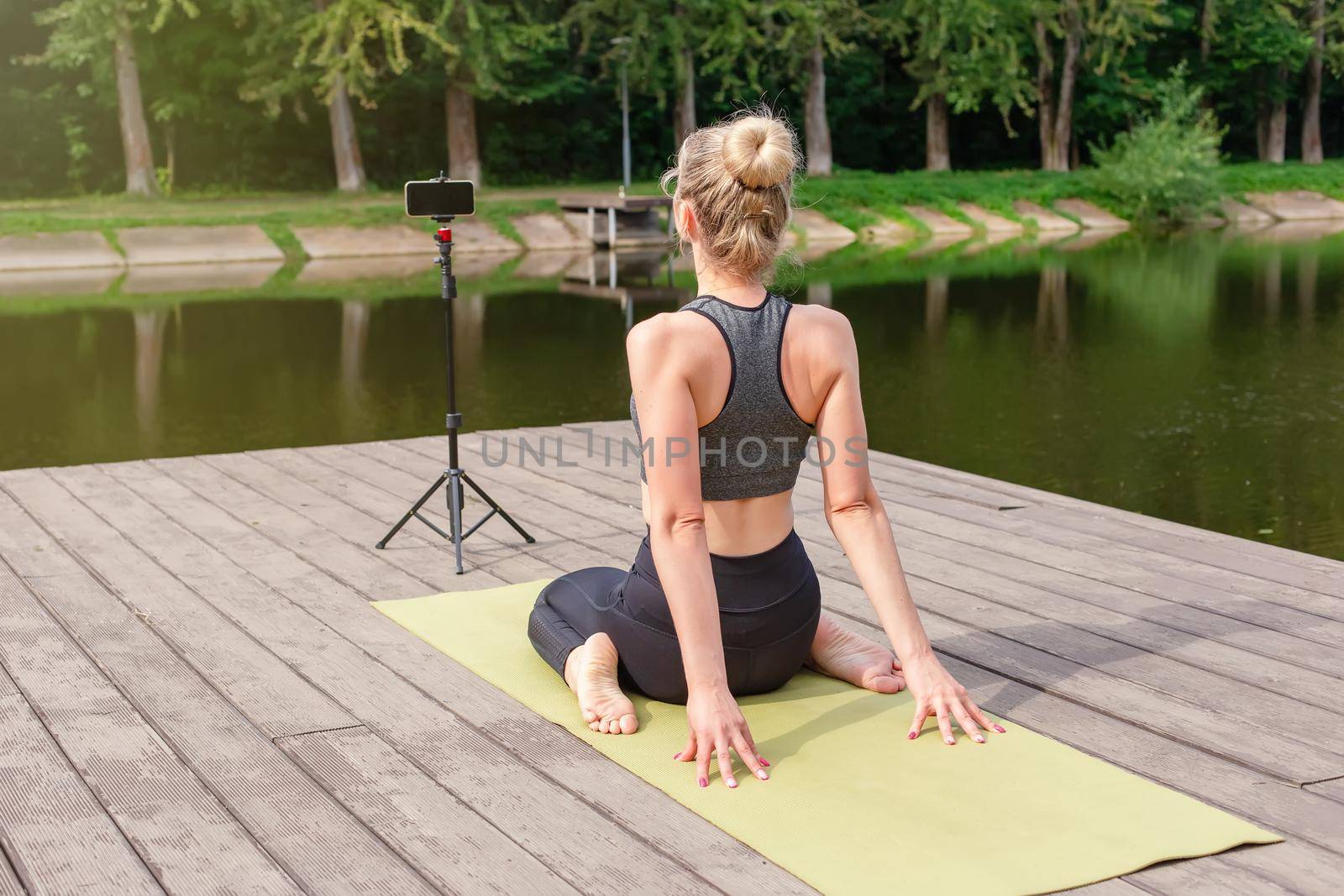 A woman in a gray top and leggings, sits on a wooden platform by a pond in the park in summer, does yoga, performs exercises, on a sports mat, in front of her smartphone on a tripod. Vertical