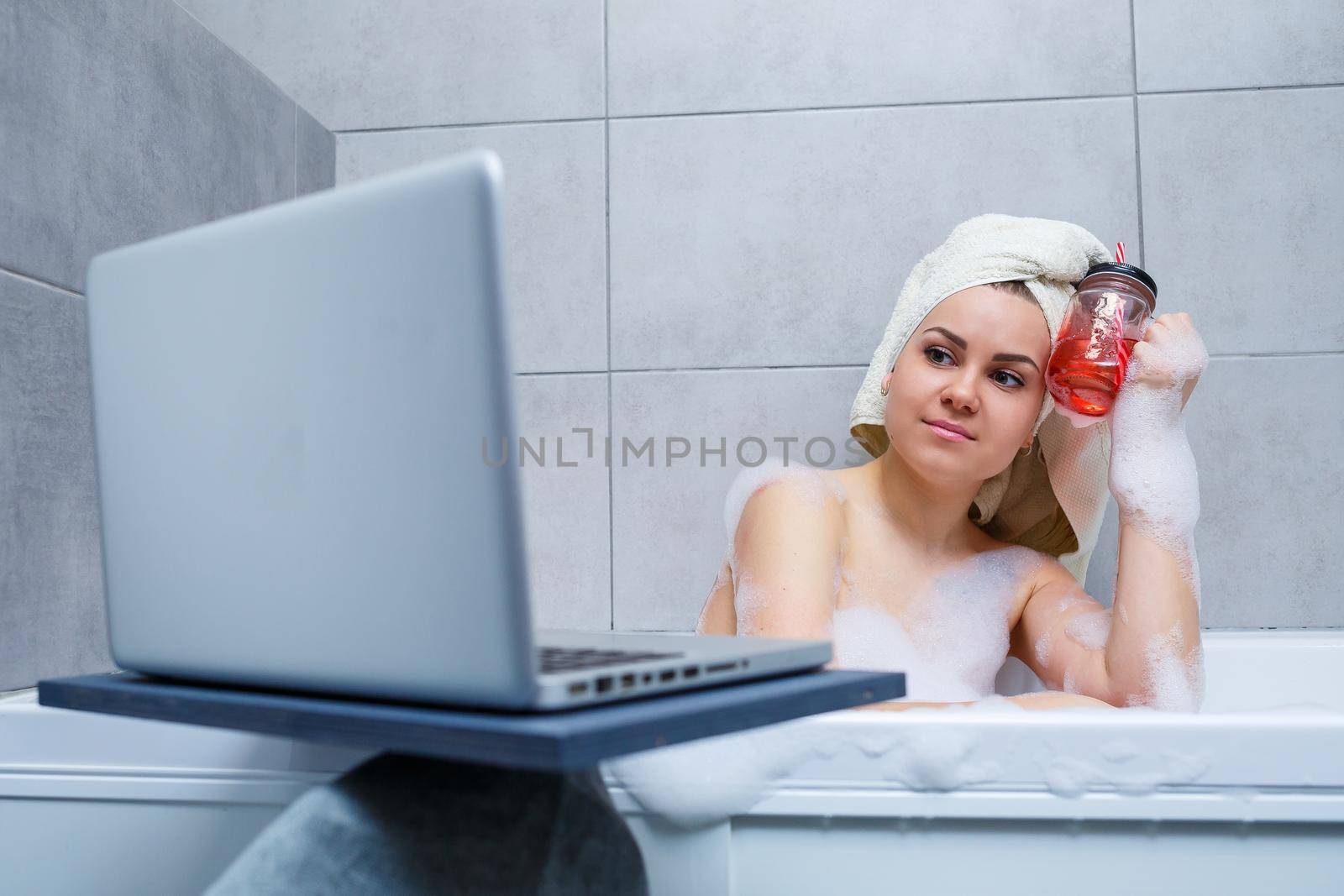 A young woman with a towel on her head drinks juice watching a movie on a laptop while sitting in a bath in a beauty salon. Relax in the bathroom without linen. Body care and relaxation concept.