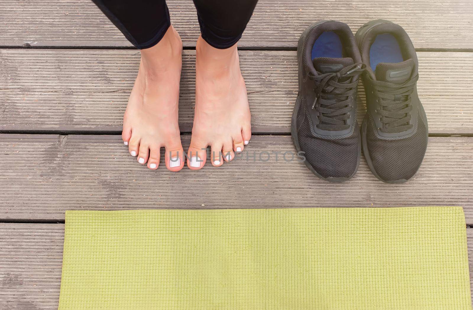 Close-up of bare feet on a wooden floor near a green sports mat, there are black sneakers nearby by Zakharova
