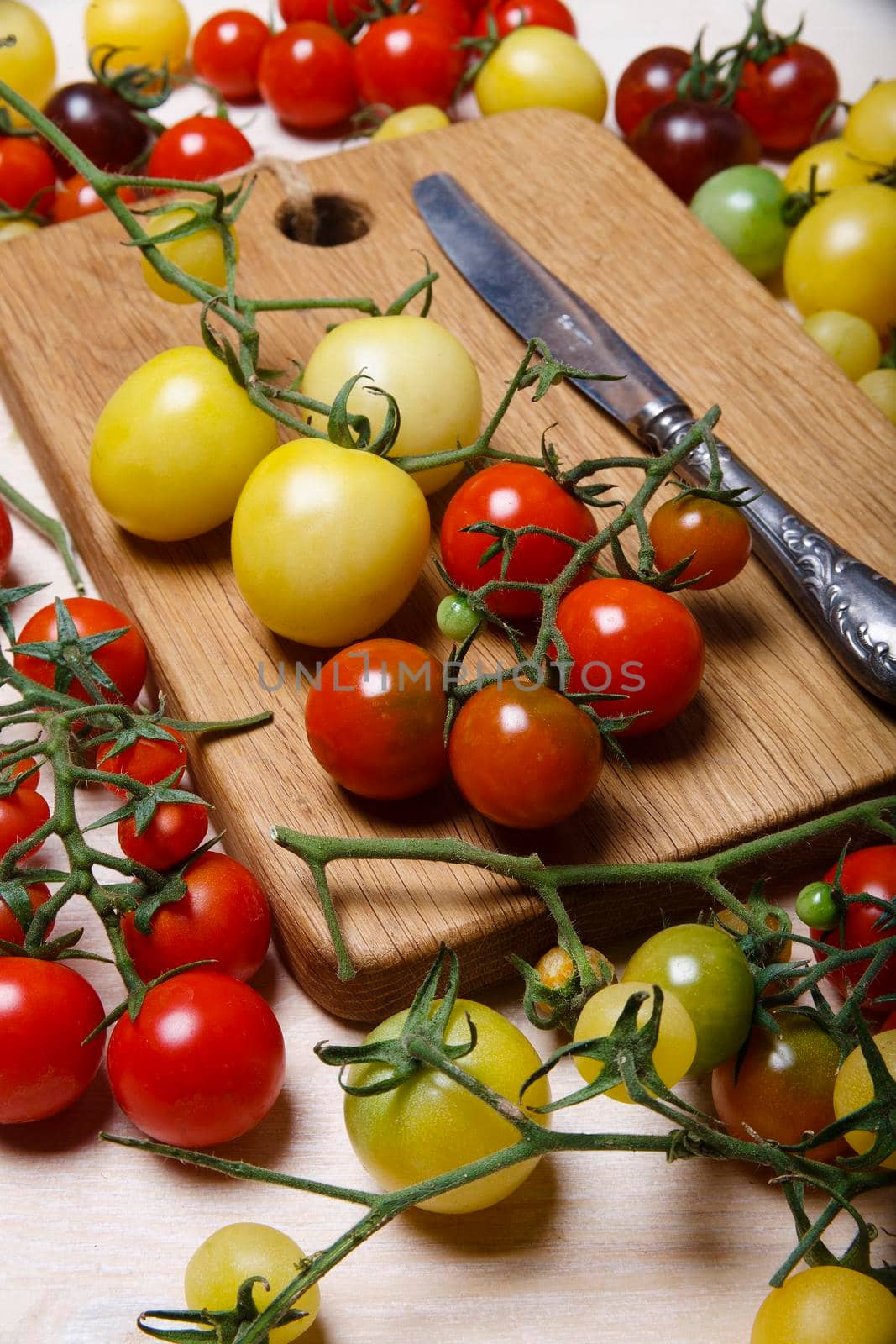 Little red, yellow, green and black cherry tomatoes on white table with wooden cutting board, nature background. by Vera_FoodandGarden