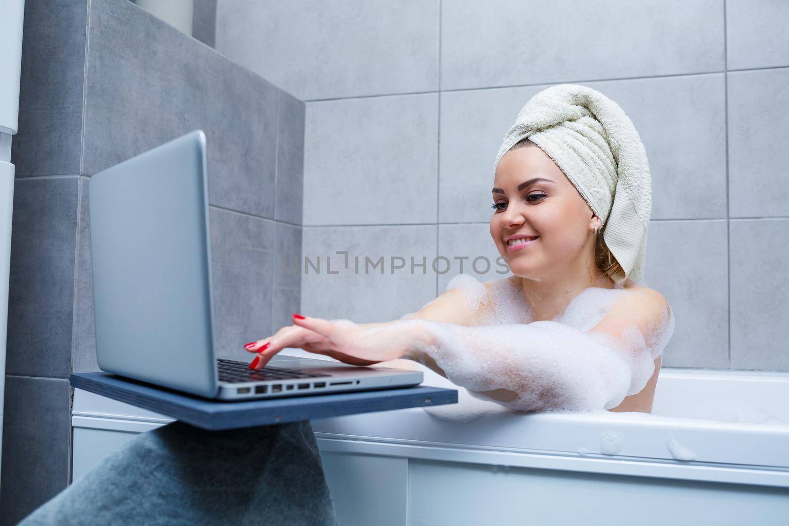 A young woman with a towel on her head is watching a movie on a laptop while sitting in a bath in a beauty salon. Relax in the bathroom without linen. Body care and relaxation concept.