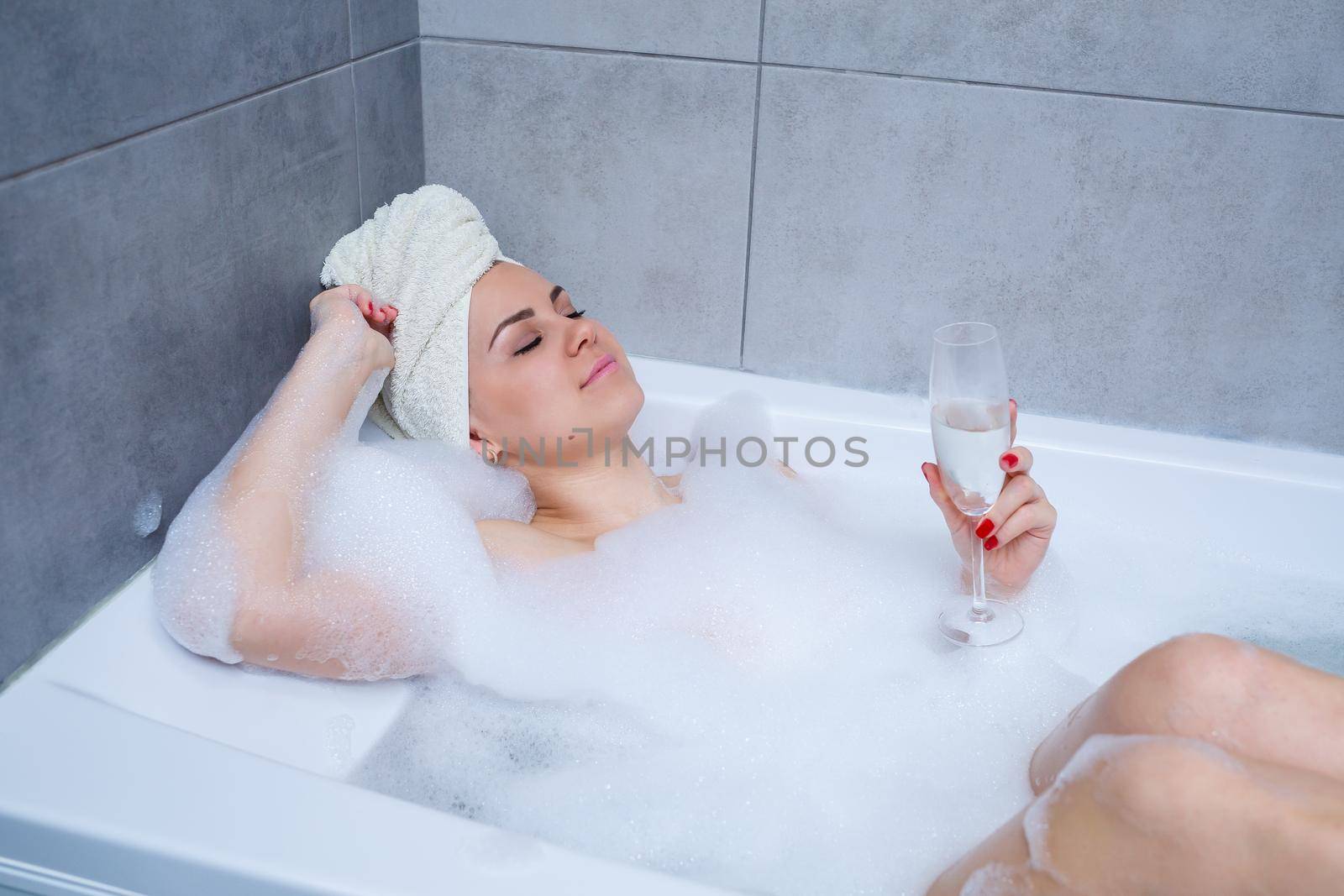 Girl woman with a white towel on her head lies with a glass of champagne in a white bathtub. In her hands a lot of pee soap. Full whirlpool foam. Relax after a hard day. Spa relaxing procedure