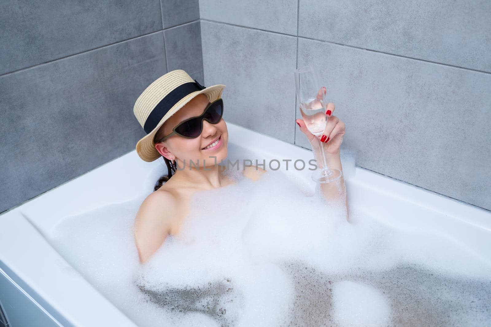 Beautiful young woman in a hat on her head and sunglasses drinking a cocktail takes a bath at home. Relax after a hard day. The spa is a relaxing procedure.