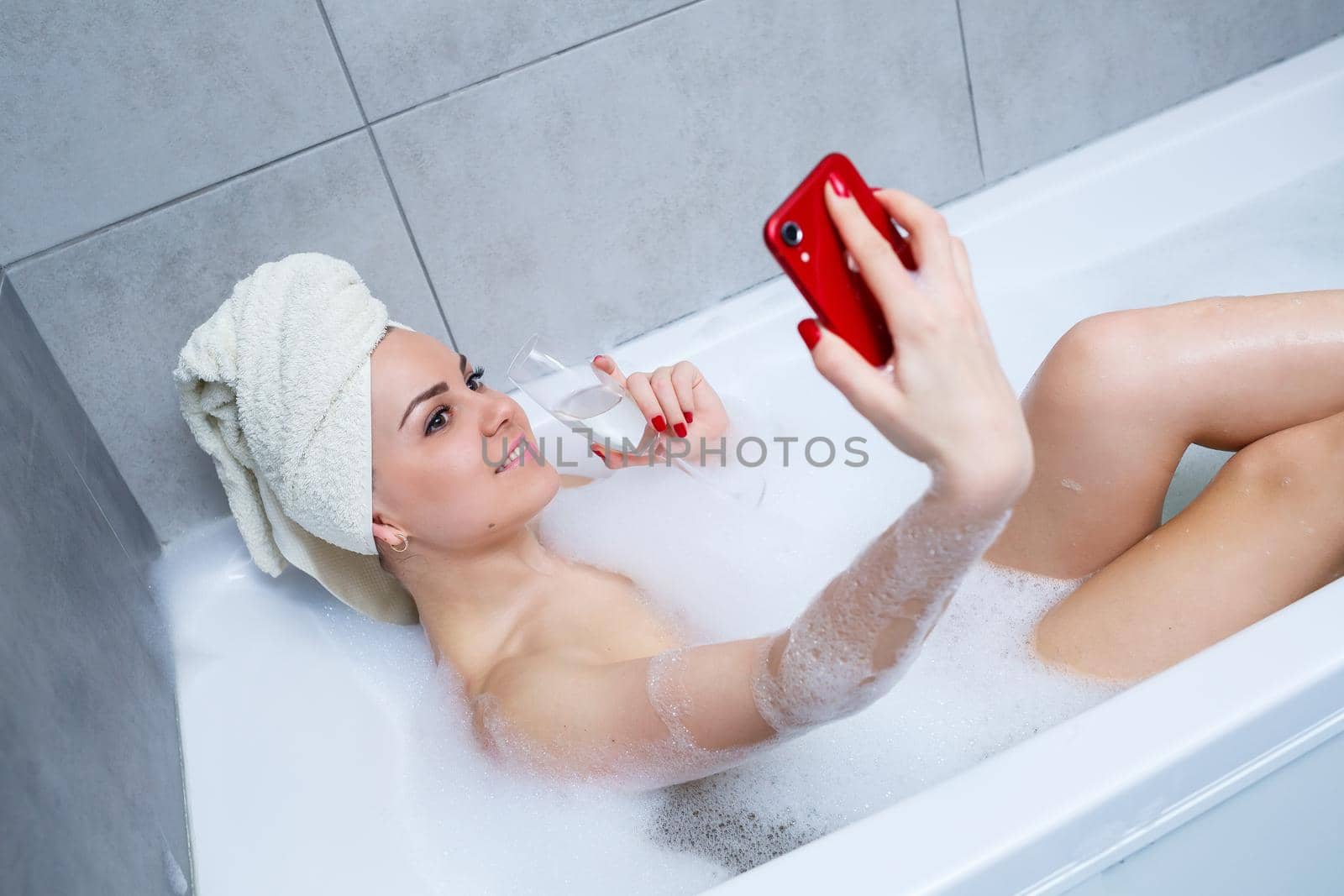 Girl woman with a towel on her head lies with a glass of champagne in a white bathtub and takes a selfie on the phone. In her hands a lot of drunk soap. Relax after a hard day. Spa relaxing procedure