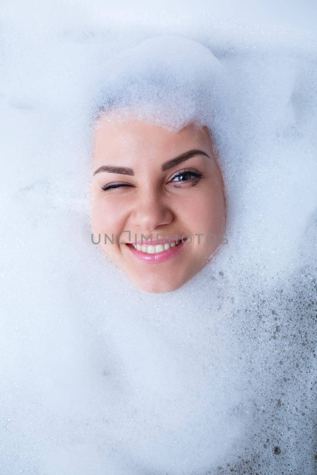 Closeup portrait of a girl in a bathtub and white foam around her face. Different emotions, grimaces, woman smile