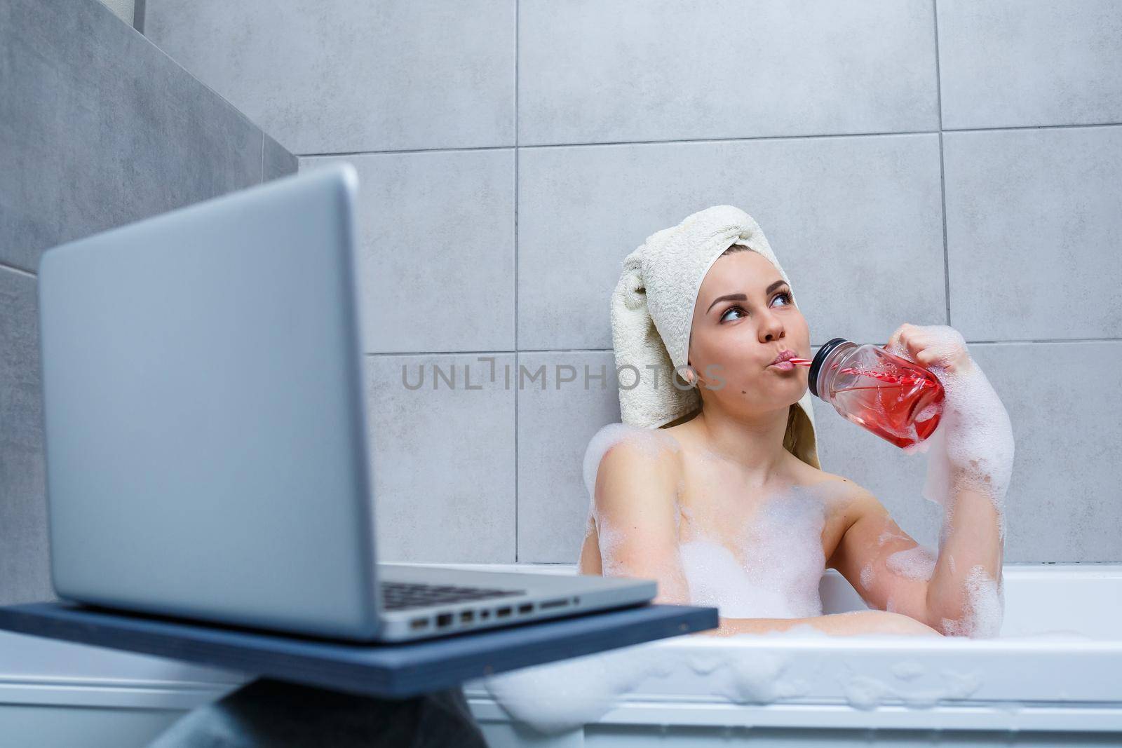 A young woman with a towel on her head drinks juice watching a movie on a laptop while sitting in a bath in a beauty salon. Relax in the bathroom without linen. Body care and relaxation concept.