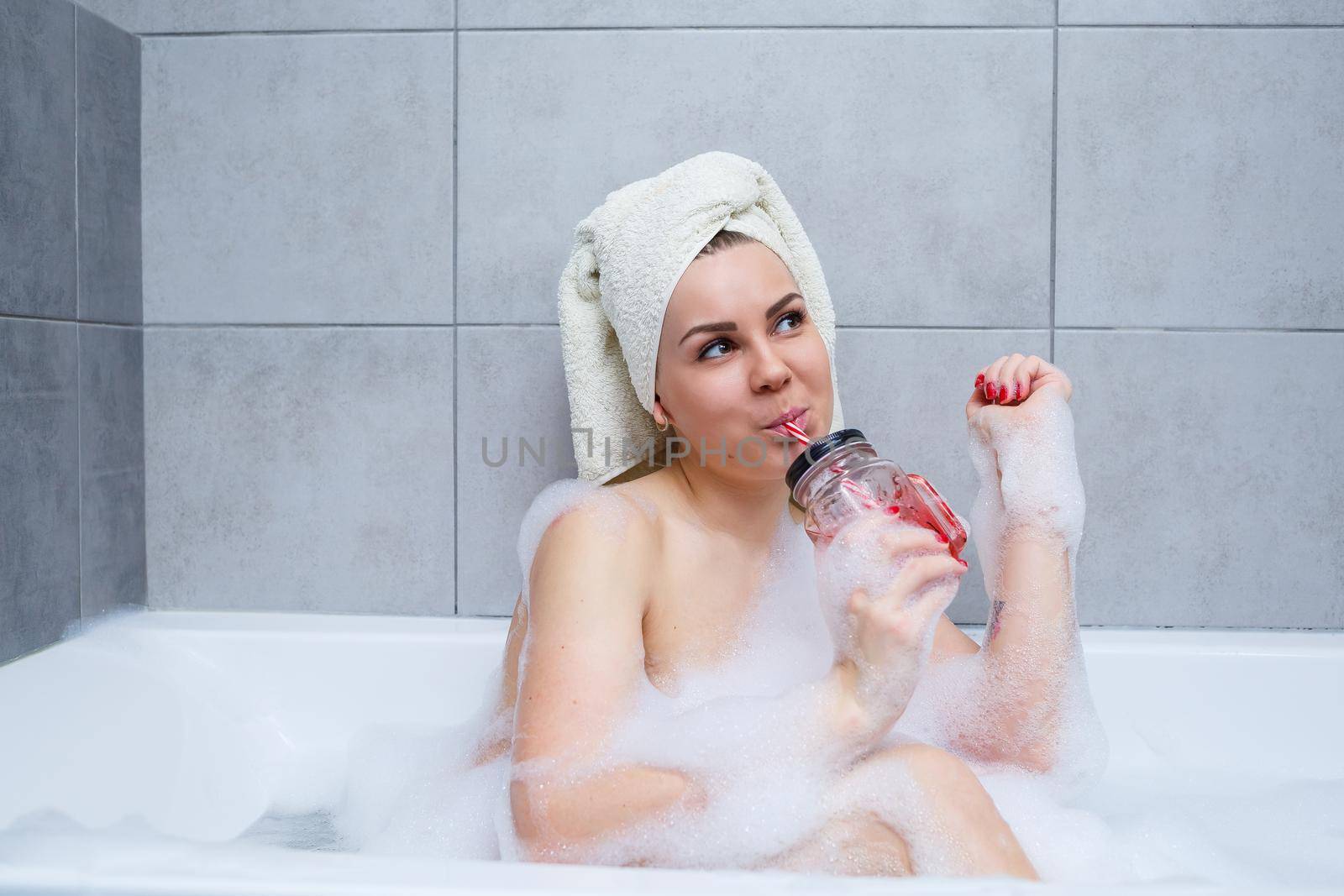 Beautiful young woman with a towel on her head drinking a cocktail takes a bath at home. Relax after a hard day. The spa is a relaxing procedure.