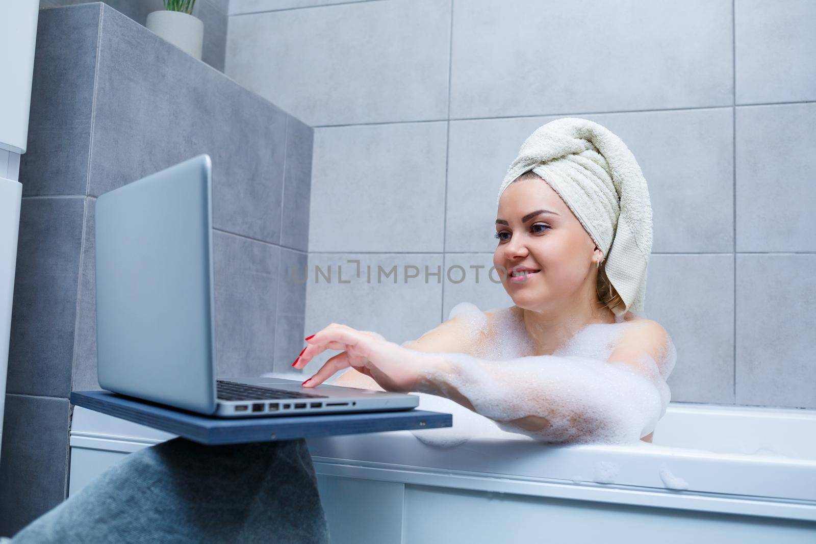 A young woman with a towel on her head is watching a movie on a laptop while sitting in a bath in a beauty salon. Relax in the bathroom without linen. Body care and relaxation concept.
