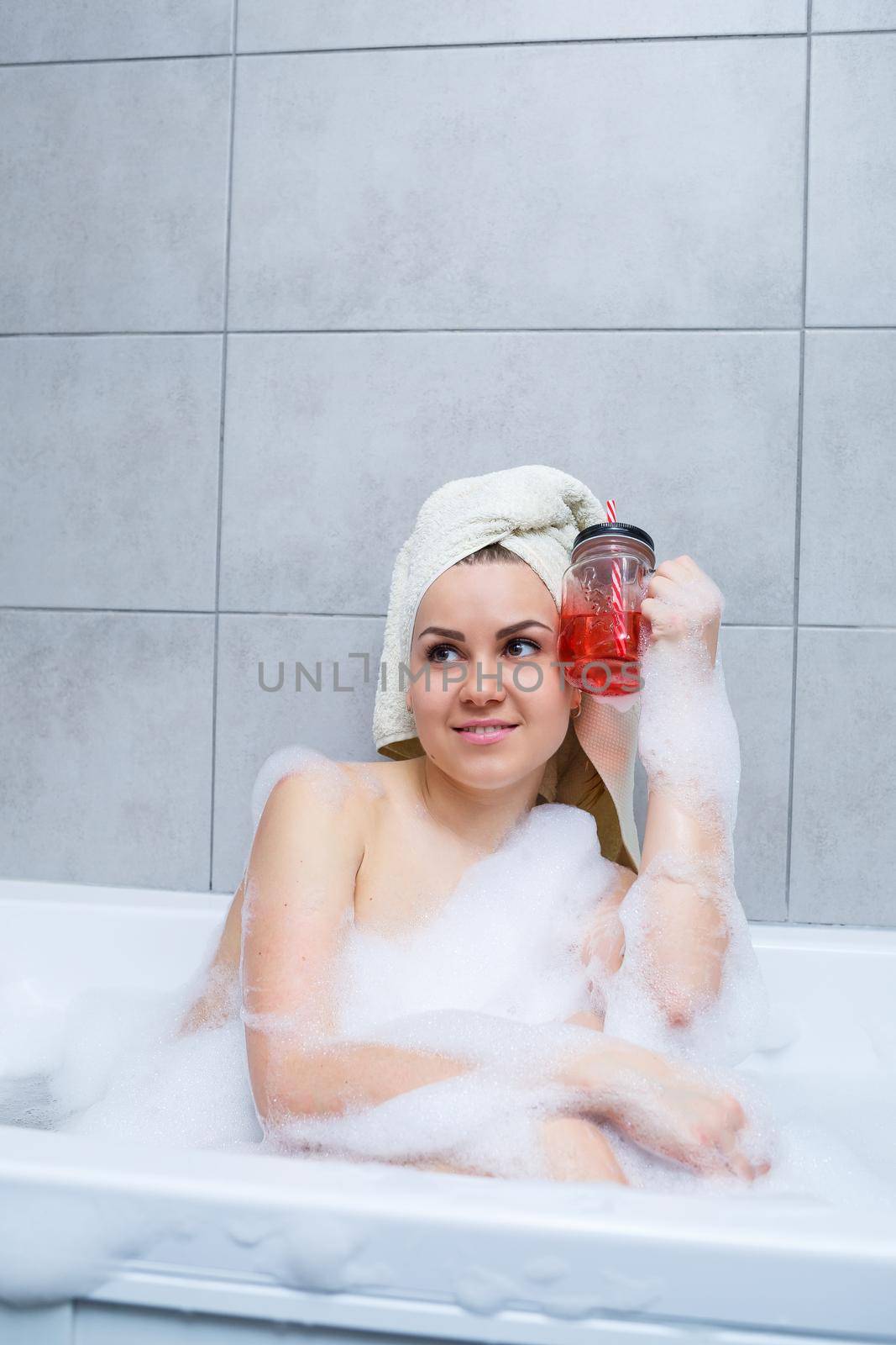 Beautiful young woman with a towel on her head drinking a cocktail takes a bath at home. Relax after a hard day. The spa is a relaxing procedure.