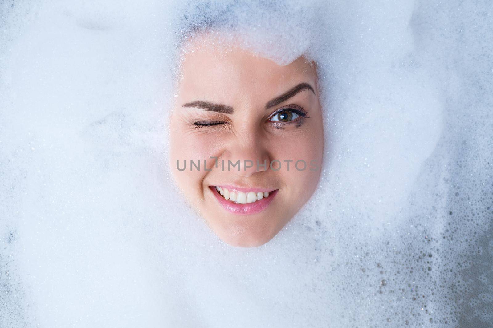 Closeup portrait of a girl in a bathtub and white foam around her face. Different emotions, grimaces, woman smile
