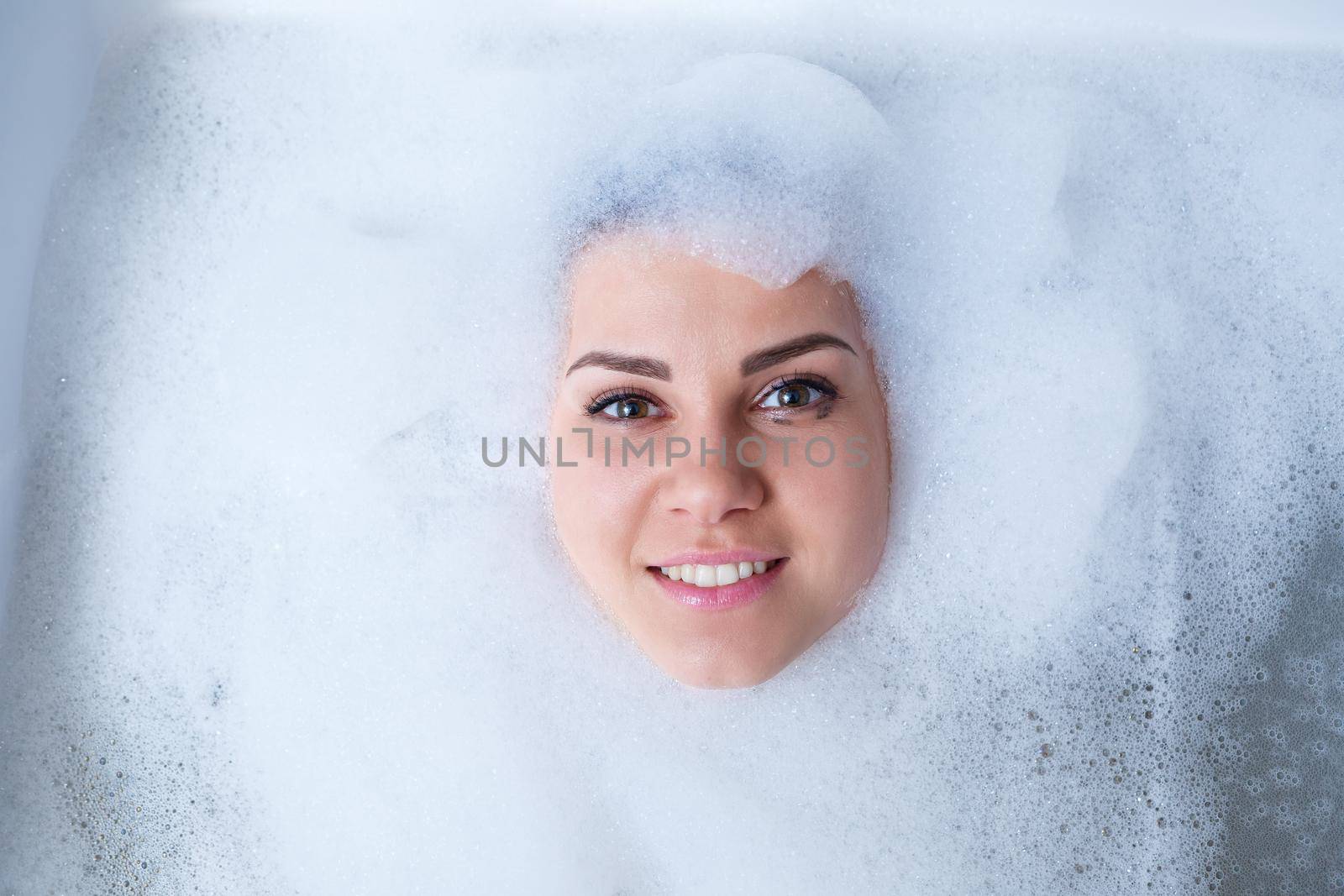 Closeup portrait of a girl in a bathtub and white foam around her face. Different emotions, grimaces, woman smile