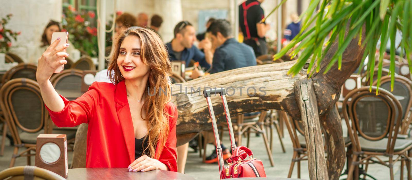 Tourist young caucasian woman in a red jacket with suitcase takes a selfie at the table in cafe outdoors