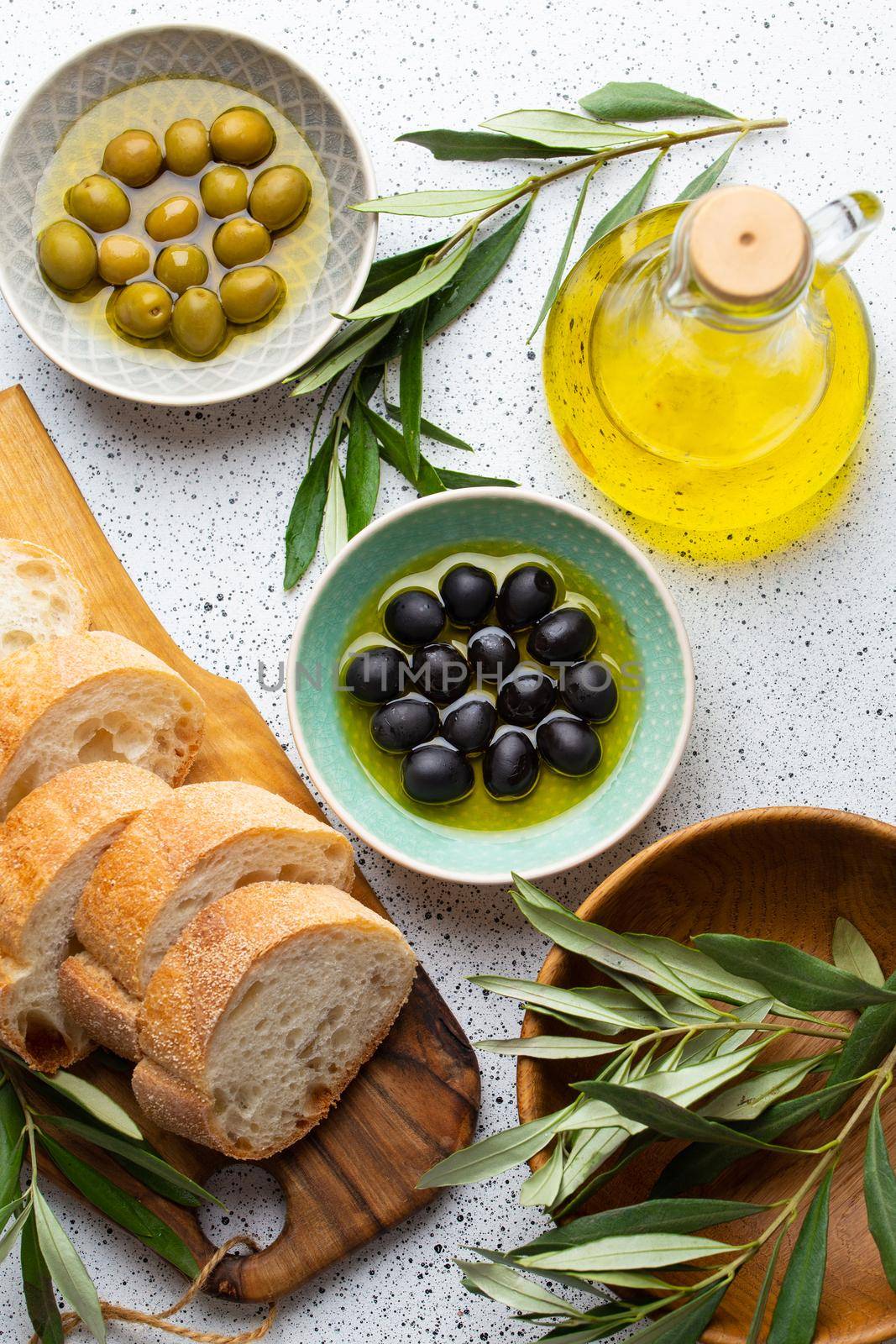 Green and black olives with olive oil in a glass bottle, olive tree sprigs and cut fresh ciabatta bread on wooden cutting board. White rustic background, mediterranean food concept, top view